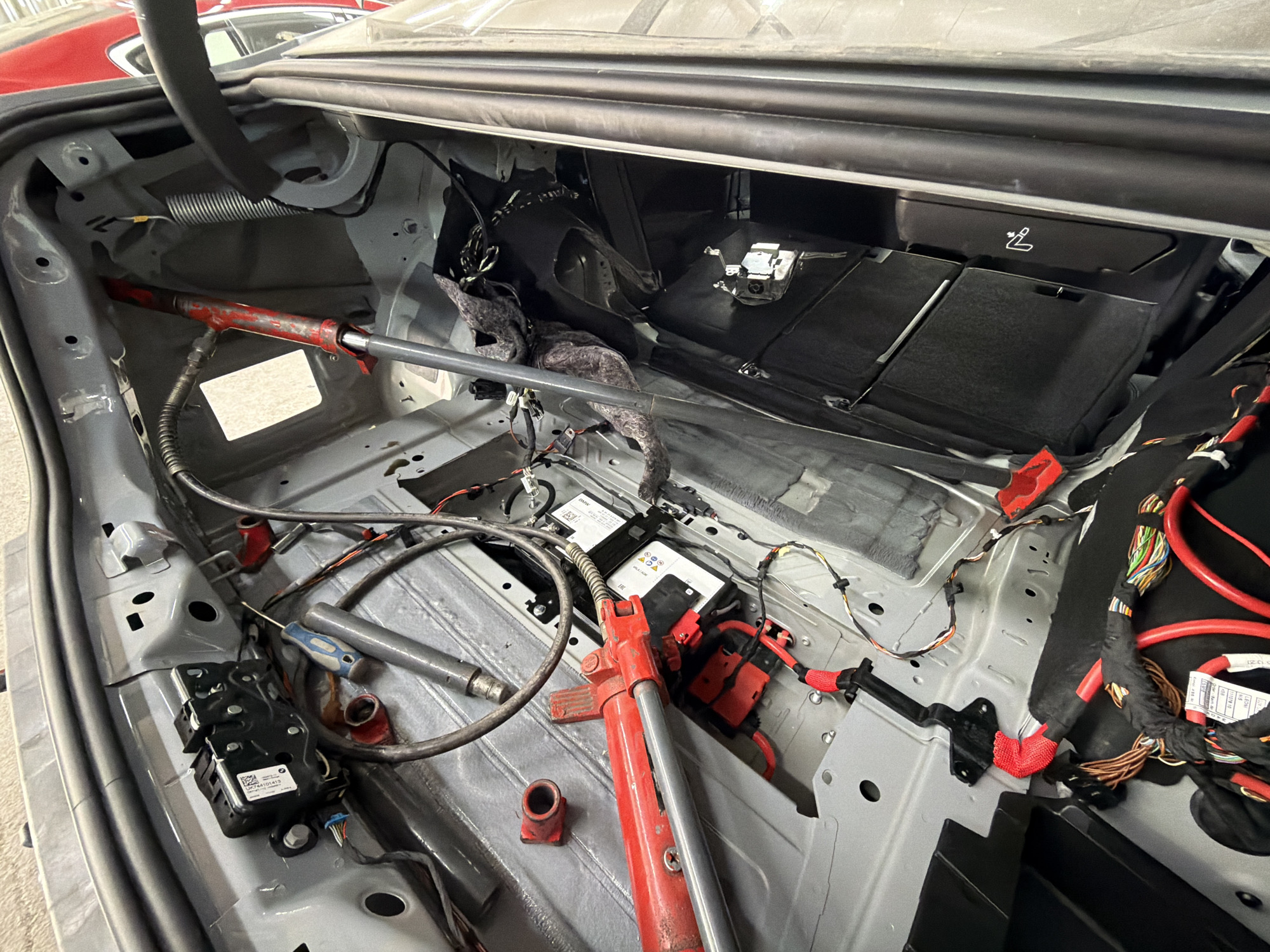 The image shows the stripped-down interior of a car trunk. The trunk's interior lining and components have been removed, exposing the vehicle's frame, wiring, and mechanical parts. Several tools are visible, including a jack and a screwdriver. Various electrical components and wires are scattered throughout the trunk space. The scene appears to be in a garage or workshop setting. Burgas, Bulgaria is not discernible in the image.