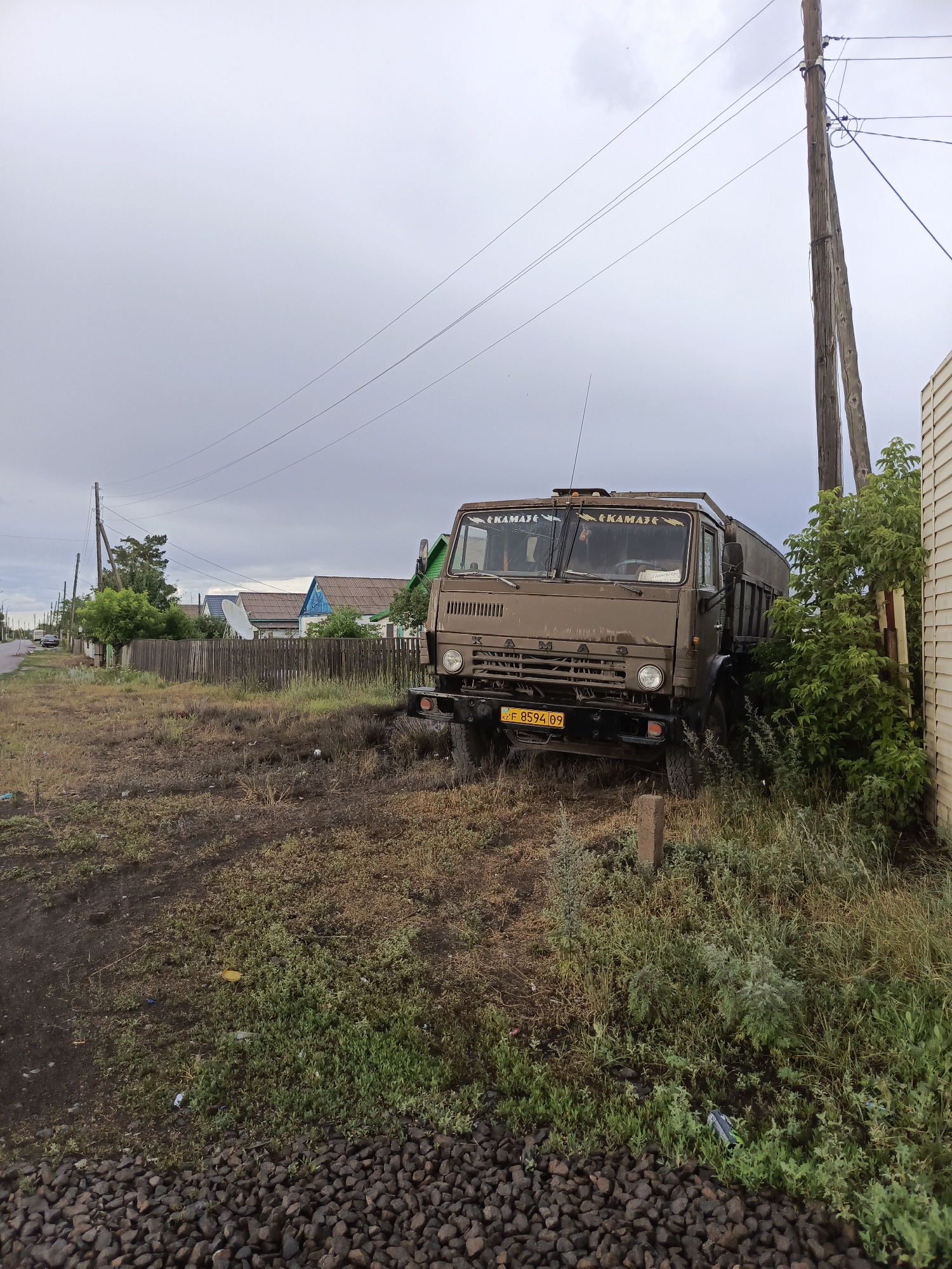 This image depicts a dusty, rural roadside scene in Shakhtinsk, Kazakhstan. The main subject is a weathered, brown KAMAZ truck parked precariously close to a wooden building and an overgrown bush. The truck's windshield has two stickers with the KAMAZ logo. A yellow license plate is visible on the front bumper, displaying the characters "KZ F 8594 09".
The setting appears to be a residential area, with a wooden fence running along the properties. Several houses with colorful roofs are visible in the background, along with trees and a satellite dish. A power line pole with multiple wires runs vertically along the right side of the frame, stretching into the cloudy sky. The ground is covered in dry grass, dirt, and small rocks. Some litter is scattered on the ground.
The sky is overcast with heavy gray clouds, suggesting a possibility of rain or a general dull day. The time of day is likely late afternoon or early evening, given the subdued lighting. There are no people visible in the frame. The overall mood is one of stillness and perhaps neglect, with the truck appearing to be out of commission or temporarily parked. The environment suggests a relatively remote or less developed area.