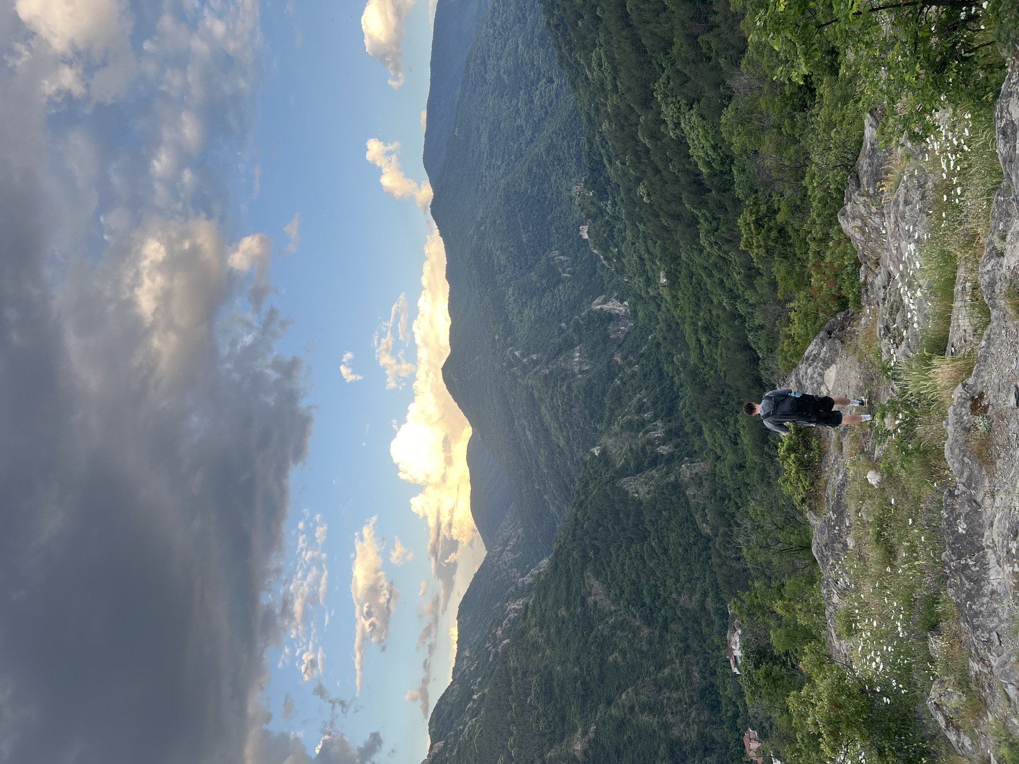 The image captures a scenic view from what appears to be a rocky cliff edge. A person with a backpack is standing on the cliff, facing toward the mountainous landscape. Lush green forests cover the mountains in the background, under a sky that is partially cloudy with both bright and dark clouds. The landscape suggests a natural environment.