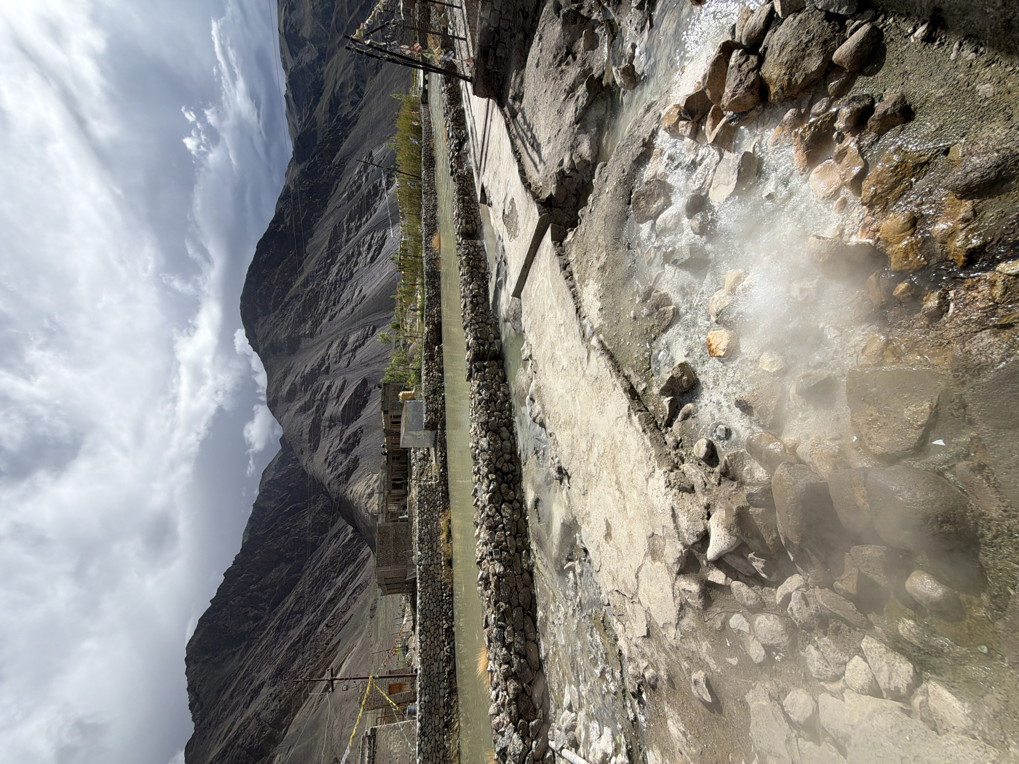 The image shows the Chumathang hot springs in India, a geothermal area located in the Ladakh region. The scene is dominated by a natural landscape of rugged mountains and a river. The hot springs are visible in the foreground, with steam rising from the water and surrounding rocks. The water is a cloudy, greenish-brown color. Concrete pathways and stone walls have been constructed around the springs, likely for visitor access and to manage the flow of water. In the background, there are buildings that appear to be part of a small settlement, with stone walls and simple structures. Prayer flags, a common sight in Buddhist regions, are strung between poles, adding a cultural element to the scene. The sky is partly cloudy, with dramatic cloud formations overhead, suggesting variable weather conditions. There are no people visible in the image, nor any obvious actions or interactions taking place. The overall impression is of a natural, yet managed, geothermal site within a remote mountain environment.
