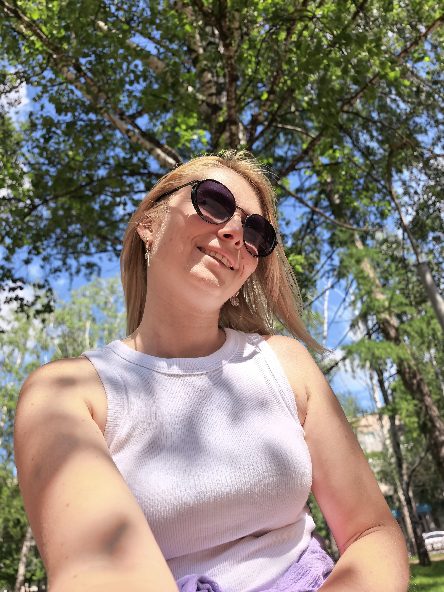 The image shows a blonde woman with sunglasses and a white tank top outdoors, looking up towards the sky. She is smiling and appears to be taking a selfie. Green trees and blue sky are visible in the background, suggesting a park or outdoor recreational area. There are no specific landmarks visible that would indicate a precise location within Sterlitamak, Russia. The lighting suggests it is daytime, possibly late morning or afternoon, with sunlight filtering through the trees, casting dappled shadows on the woman's arm and clothing.