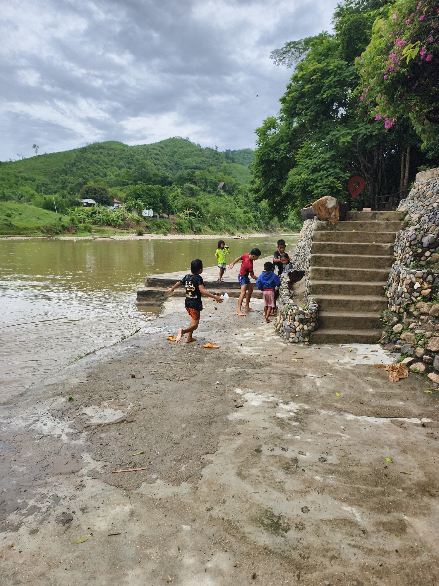 The image shows a riverbank scene with several children. Five children are present near a set of concrete stairs with stone sides leading upwards into vegetation. One child is running along the concrete platform towards the river. Another child in a red shirt and blue shorts stands on the edge of the platform near the stairs. A third child in a blue jacket and pink shorts stands between the stairs and the child in red. Two additional children are positioned near the base of the stairs. The river, murky brown, is adjacent to the platform. Beyond the river, a green, hilly landscape is visible under a cloudy sky. Houses can be seen on the hillside. The location is Unknown, Unknown.