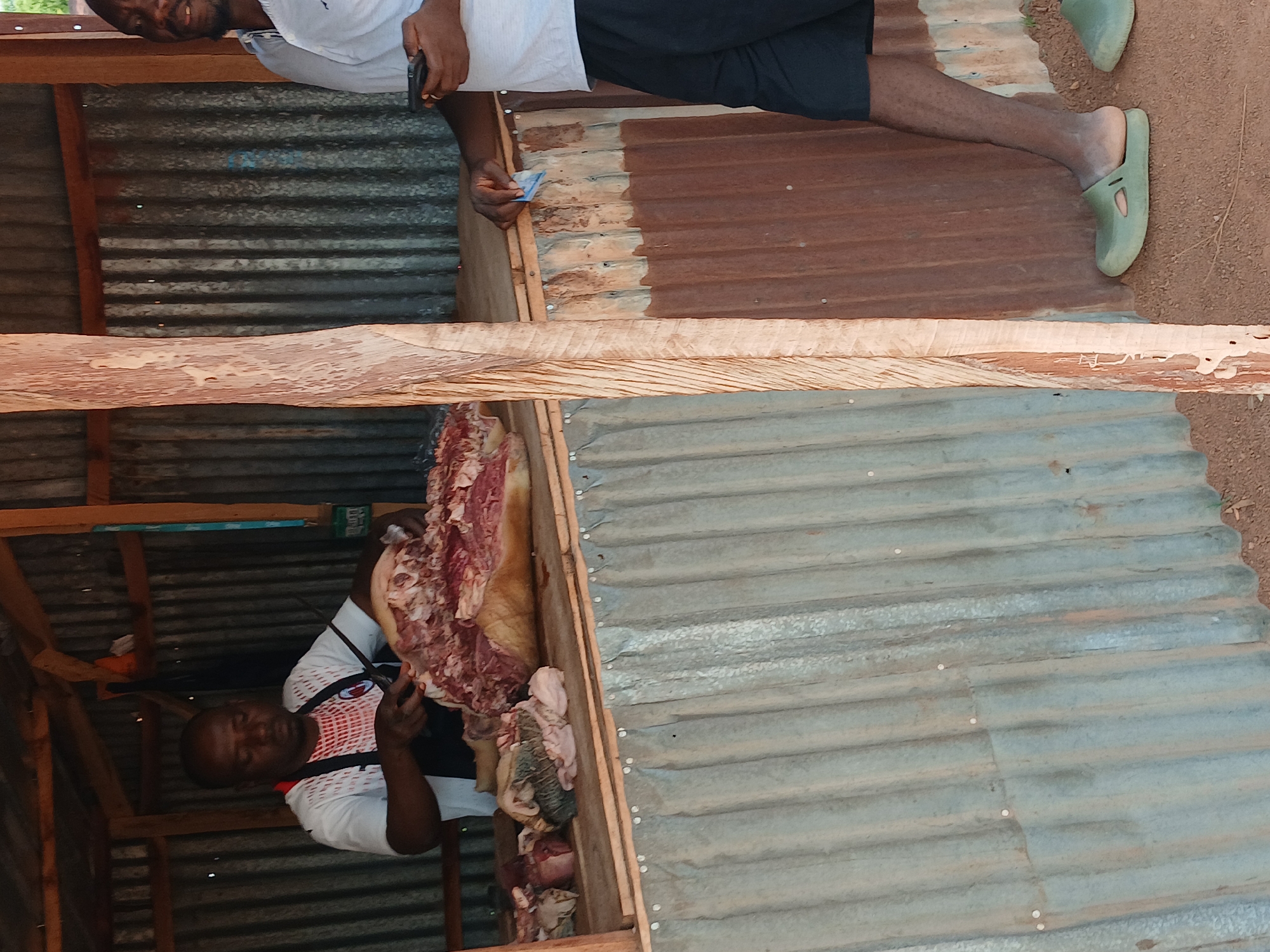 This image depicts a butcher shop in Kwang, Nigeria. Two men are present. The man on the left, wearing a white t-shirt and an apron, is actively butchering a large cut of meat, possibly pork, with a knife. He is positioned behind a wooden counter laden with various cuts of meat. To the right, another man, wearing a white t-shirt and black shorts, stands holding a mobile phone. The shop appears to be an open-air structure with corrugated metal walls and a wooden roof. The ground is covered with dirt. The scene suggests a commercial transaction or preparation for one.