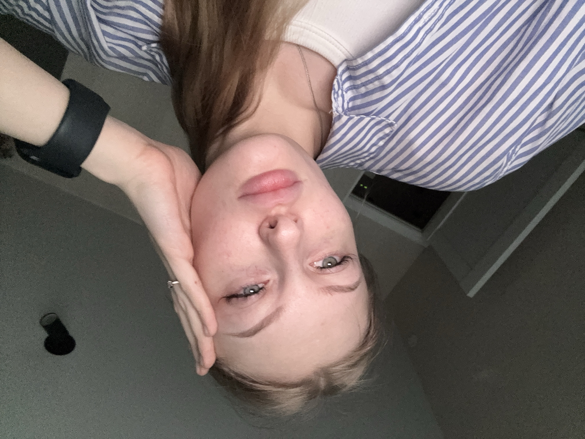 A person is holding their face with their hand, looking at the camera. They are wearing a blue and white striped shirt over a white top. They are wearing a black wristband on their left wrist. There is a ring on the ring finger of their left hand. There is a window in the background and a black object on the wall. There is no evidence to identify the location as Perm, Russia.