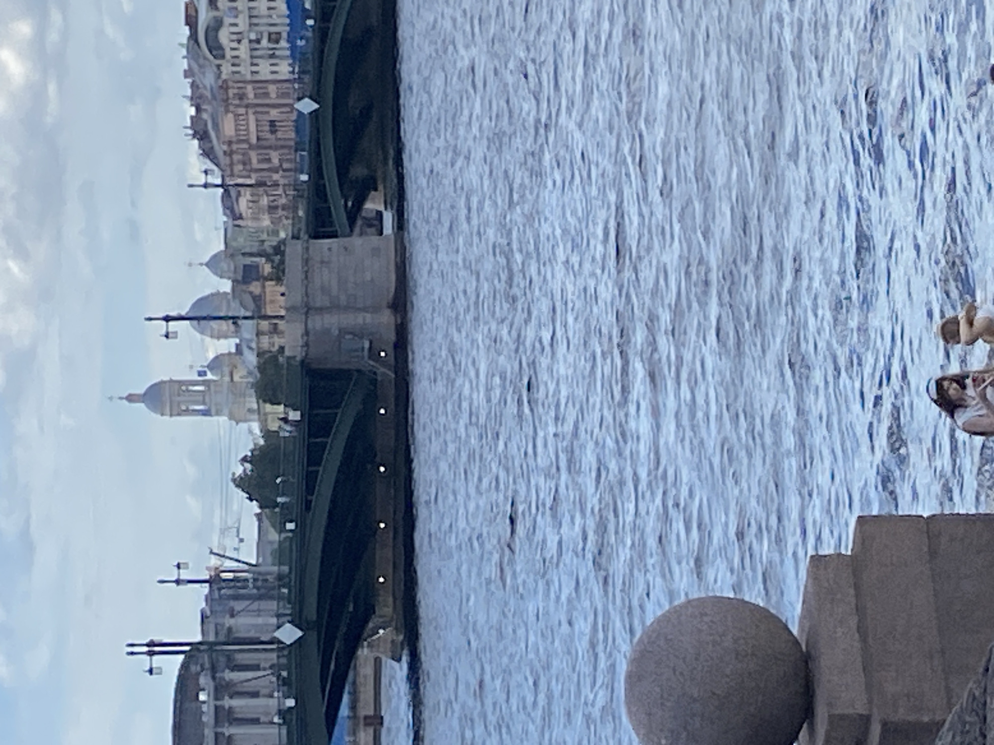 In Saint Petersburg, Russia, a wide blue river dominates the foreground, with gentle waves rippling across its surface. On the left, a large, weathered stone sphere rests atop a multi-tiered stone pedestal, indicating a waterfront promenade or park area. In the middle ground, a massive, dark green bridge arches over the river, its supports a mixture of stone and concrete. Beyond the bridge, the Saint Petersburg skyline unfolds, featuring numerous buildings with classical architecture, domes, and spires. The sky is overcast with soft clouds, suggesting it is either early morning or late afternoon. In the lower right corner, two individuals are visible near the water's edge, appearing to be seated or lounging. One person is wearing a white top and a headscarf, while the other, partially obscured, is wearing a white swimsuit. The overall scene captures a tranquil urban landscape with elements of architecture and nature.