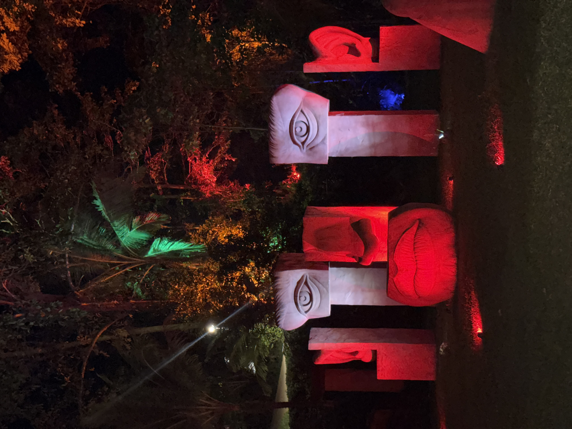 The image depicts a night scene in Ko Samui District, Thailand, featuring an outdoor art installation illuminated by colorful lights. The installation consists of several large, abstract sculptures resembling parts of human faces. In the foreground, a large, red, mouth-shaped sculpture is prominently displayed, with its lips parted. To the left and right of the mouth are vertical, white sculptures, each featuring a carved eye with an eyelid and iris. Further to the sides, there are additional vertical sculptures, one on the left with a red glow, and one on the right that appears to be an ear shape, also illuminated in red. The sculptures are placed on a grassy area. In the background, trees are visible, some of which are lit with green and red lights, creating a dramatic contrast against the dark sky. A faint white light from a lamppost is also visible in the upper left portion of the image. The overall impression is one of a visually striking, nighttime art display in a tropical setting.