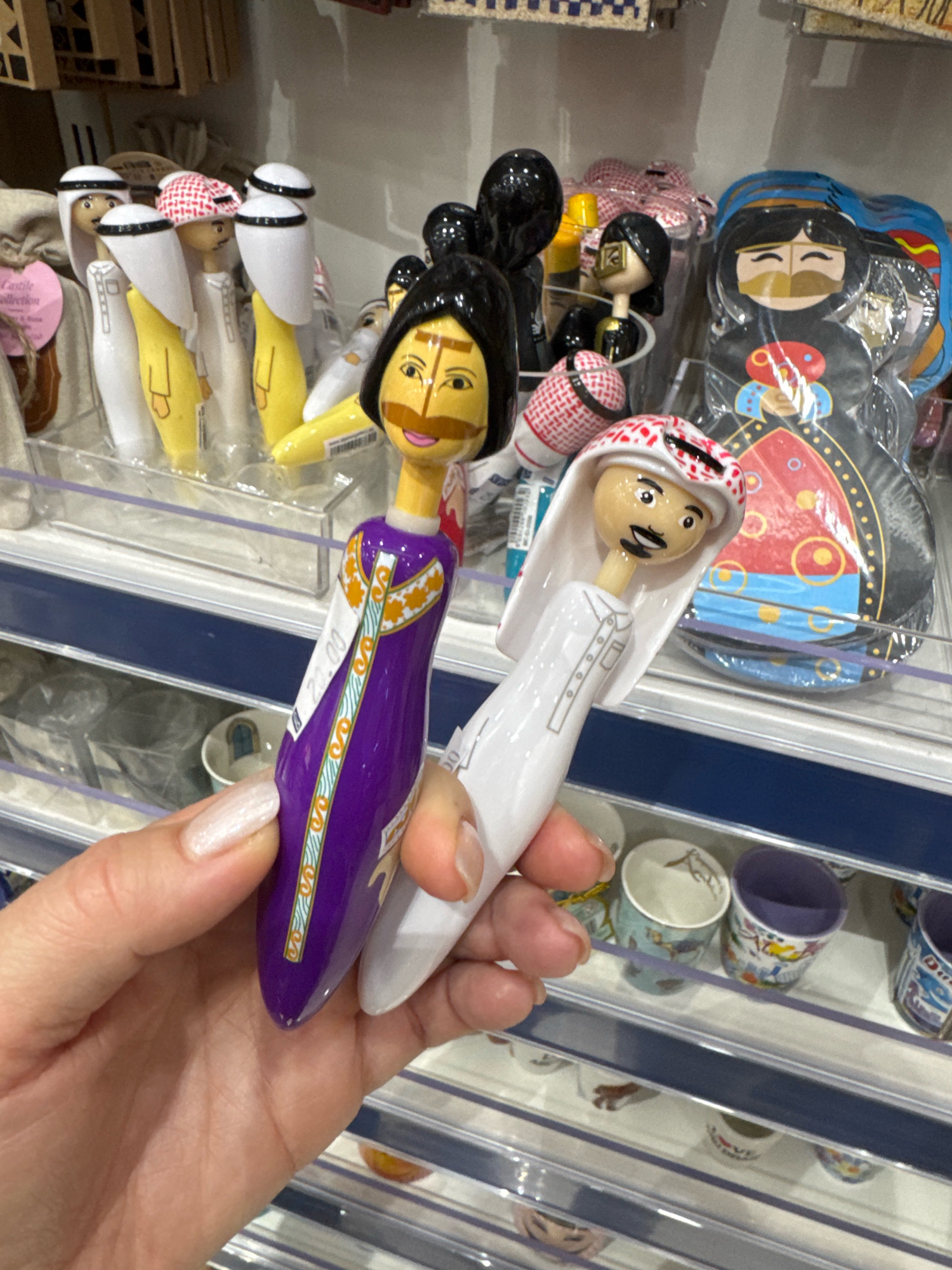 A close-up, eye-level shot captures a hand holding two novelty pens shaped like cartoonish figures dressed in traditional Middle Eastern attire. The pen in the foreground, held by the fingers and thumb, depicts a figure in a purple thobe with gold trim. The pen behind it, gripped by the thumb and index finger, shows a figure in a white thobe. Both figures have stylized facial features. The background reveals a retail setting with shelves displaying more similar novelty items, including figurines and small decorative cups. The overall impression is of a souvenir shop or gift store. The location is identified as Al Matar, United Arab Emirates, suggesting this is a retail display within that region.