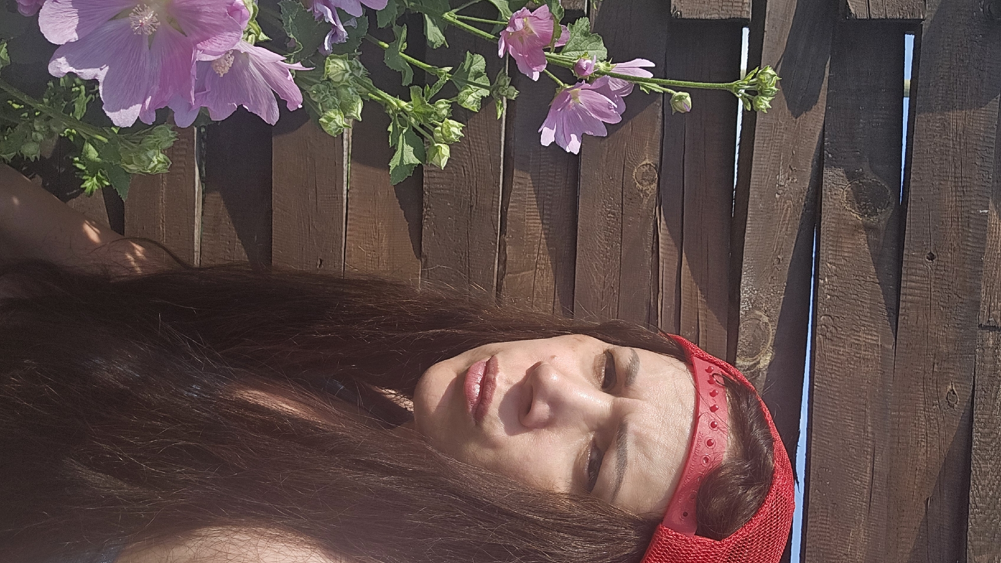 A woman with long, dark brown hair is lying down outdoors, with her head resting on a wooden surface. She is wearing a red headband. Her eyes are closed, and her lips are slightly parted, suggesting a relaxed or possibly sleeping state. She appears to be in her late twenties or thirties.
Above her, a branch of pink and purple hollyhock flowers is visible against a rustic wooden fence. The fence is made of weathered, dark brown planks, with some gaps between them allowing glimpses of a bright blue sky. Sunlight casts shadows from the flowers and the fence, indicating it is daytime, likely late morning or afternoon, with clear weather.
The scene is set outdoors, possibly in a garden or backyard, given the presence of flowers and a fence. The overall mood is peaceful and serene, enhanced by the soft lighting and the natural elements. There is no visible text in the image. The location context provided is Ermolaevo, Russia.