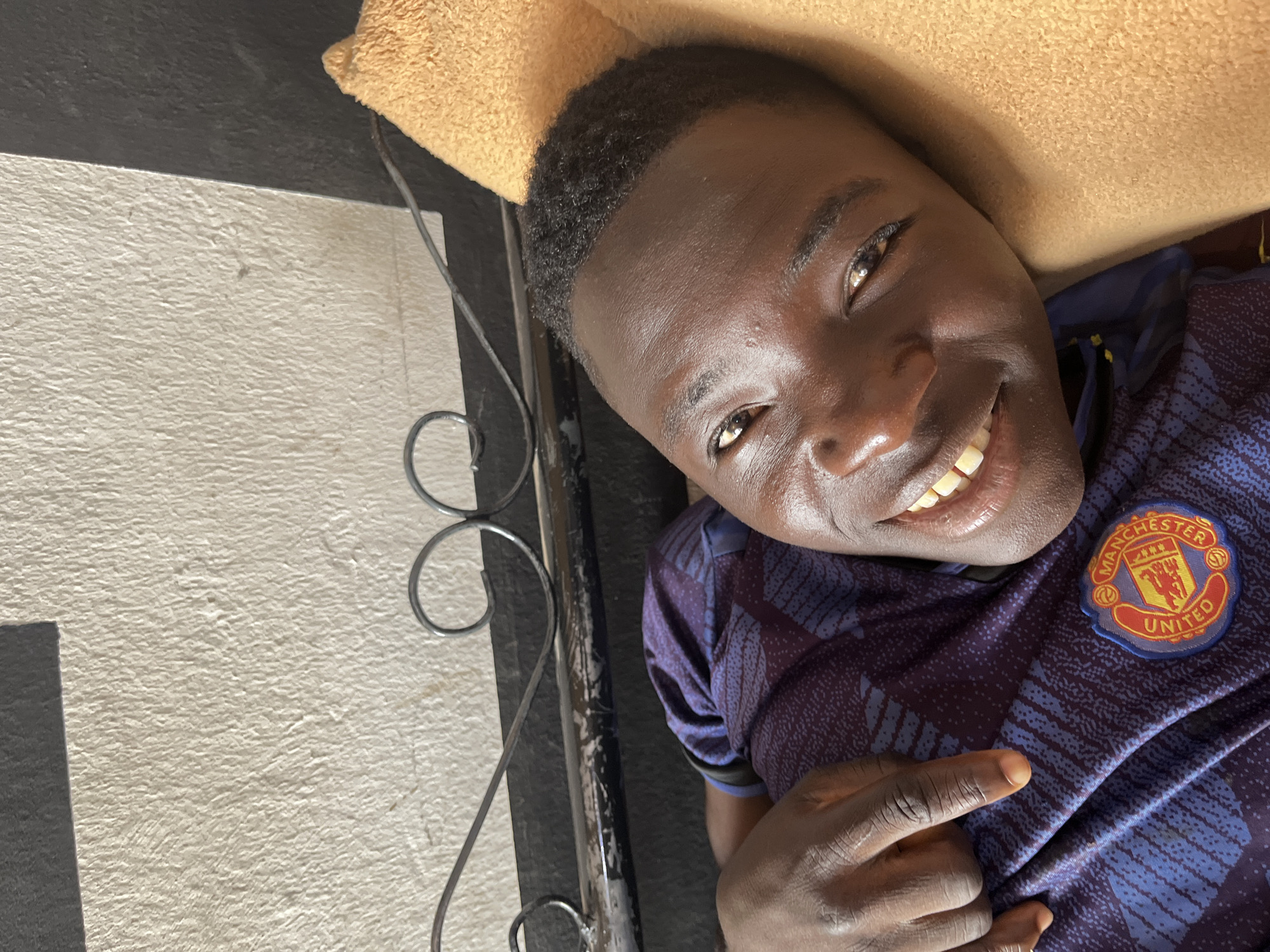 A young, dark-skinned male is lying down, looking toward the camera and smiling. He is wearing a dark blue and purple Manchester United jersey. He is positioned on a tan-colored pillow or cushion. A black, wrought-iron piece of furniture is visible to his left. The wall to his left is light-colored. The location is Jos, Nigeria.