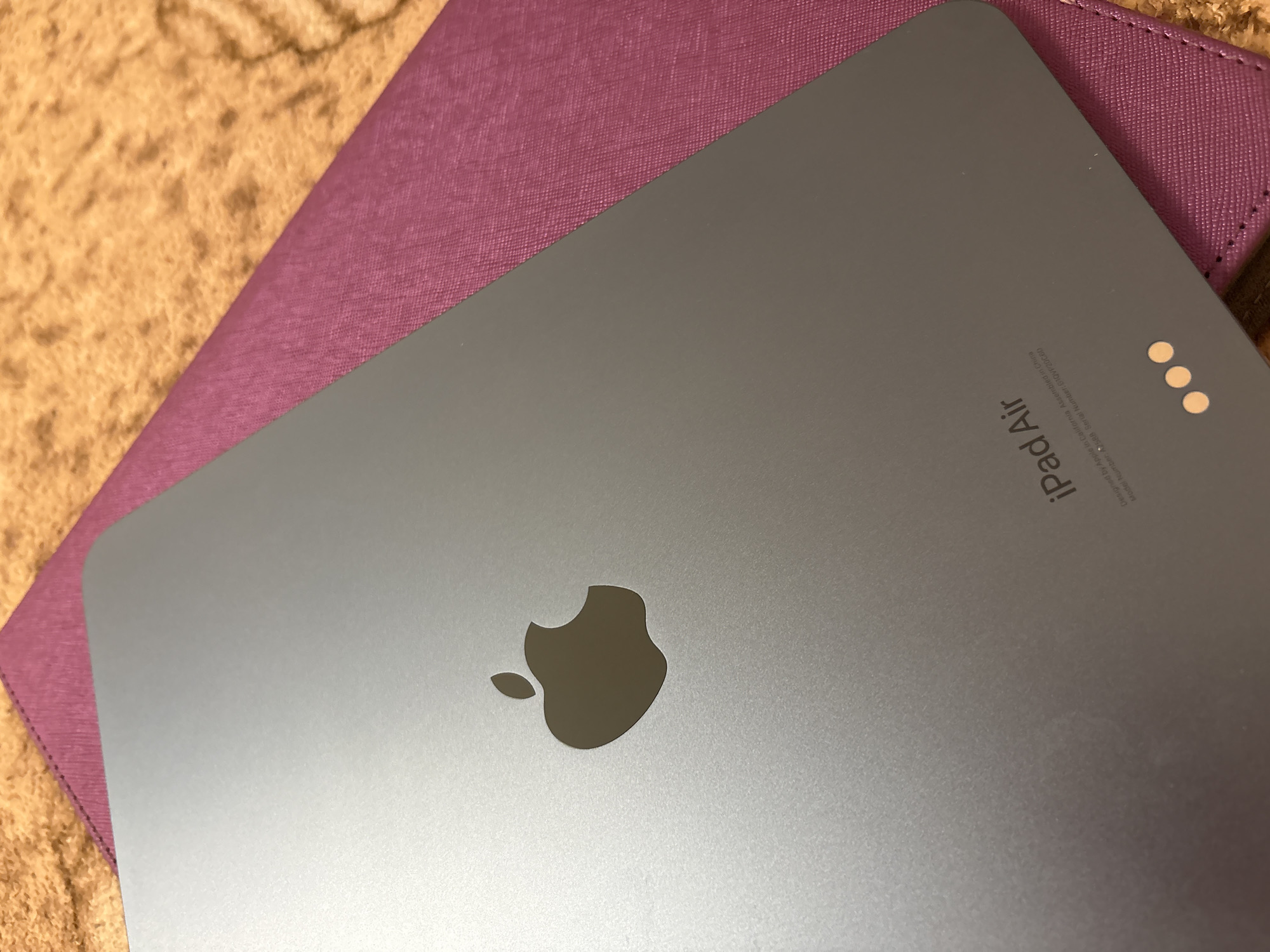 The media file shows a close-up, top-down view of the back of a gray iPad Air tablet. The iPad is resting on a textured, light brown surface, possibly a blanket or rug, and partially overlaps a reddish-purple folio or case. The Apple logo is prominently displayed in the center of the iPad's back. In the upper right corner of the iPad's back, there is small, engraved text that reads "iPad Air" and additional details about its design and manufacturing, including "Designed by Apple in California Assembled in China" and model and serial numbers. Three small, circular dots are also visible near the text. The lighting suggests an indoor setting, and there are no people or active events depicted.