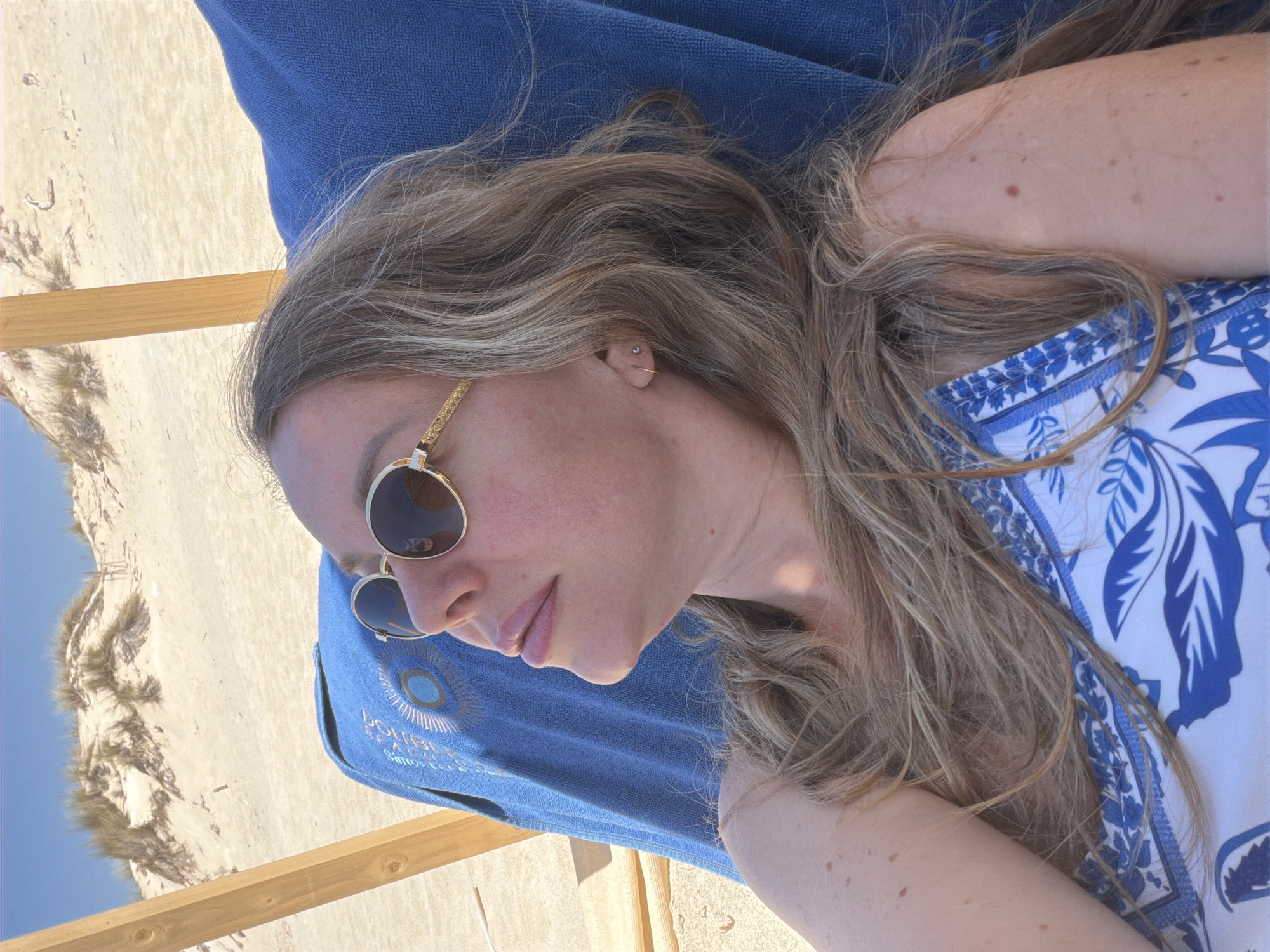A woman lies on a beach lounge chair covered with a blue towel. She wears sunglasses and a blue and white patterned top. Her hair is light brown and wavy. The background shows sand dunes under a clear blue sky.