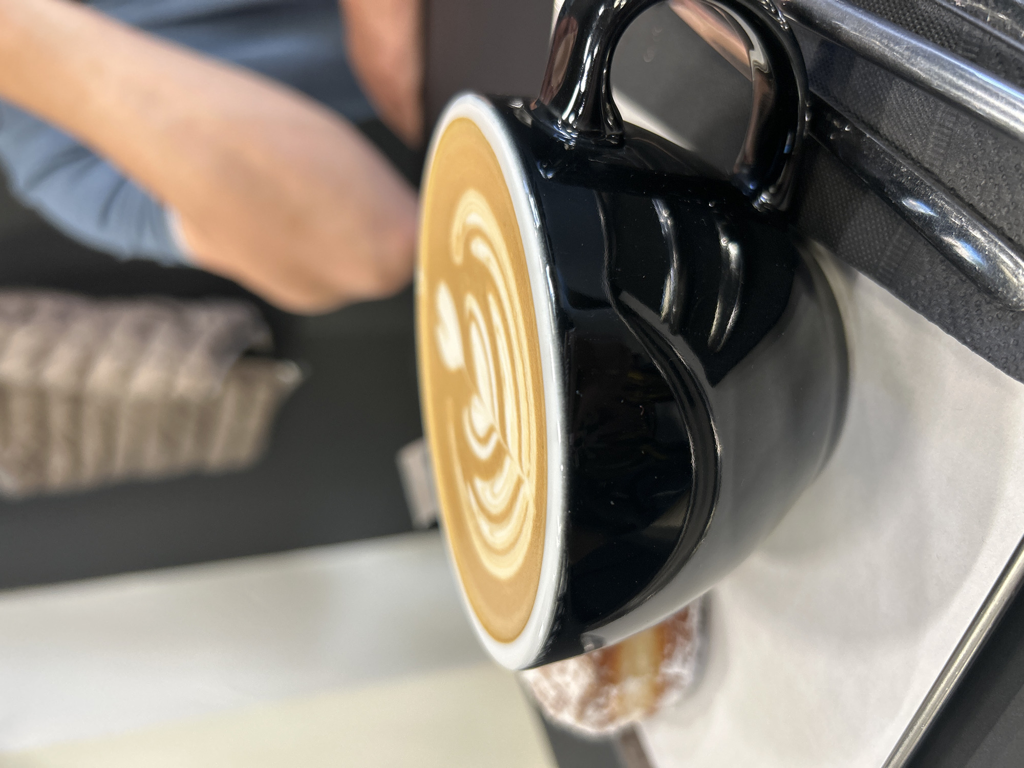 A close-up shot depicts a black ceramic mug filled with a latte, showcasing latte art. The mug rests on a white paper sheet on what appears to be a metal tray, next to a partially visible donut. In the background, a person's arm and leg are visible. The location and city of Izhevsk, Russia are not discernible from this image.