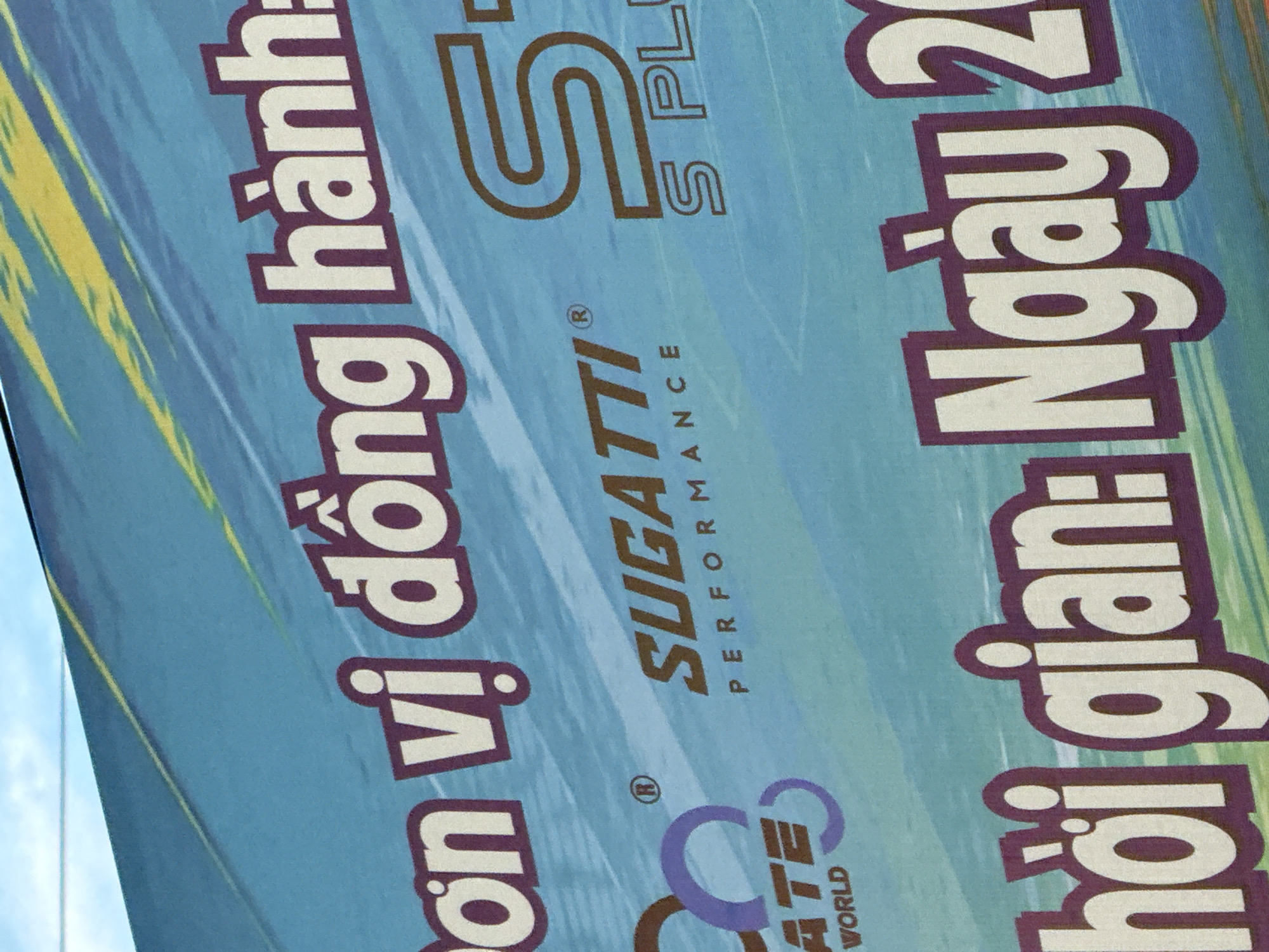 The image displays a close-up view of a large, blue banner with text and logos. The banner appears to be an advertisement or informational display, likely for an event or company. Prominently featured are large white and purple letters spelling out Vietnamese text, including "ĐỒNG HÀNH" which translates to "ACCOMPANYING" or "WALKING TOGETHER." Below this, the word "SUGATTI PERFORMANCE" is visible in a smaller, dark brown font. To the right, a stylized brown "S" logo is present, followed by "S PLUS" in a similar font. On the left side, a logo with purple rings surrounds a brown word, with "ATE WORLD" visible below. The bottom portion of the banner shows more large white and purple Vietnamese text, including "NGÀY" which means "DAY." The banner's background has a diagonal gradient of light blue to darker blue with yellow and white abstract streaks. The context suggests this banner is located in Nha Trang, Vietnam, given the Vietnamese language used. The image captures a segment of the banner, not the entire scene, so specific details about the surrounding environment, such as the city of Nha Trang itself, are not visible. There are no people or actions depicted in this specific frame.