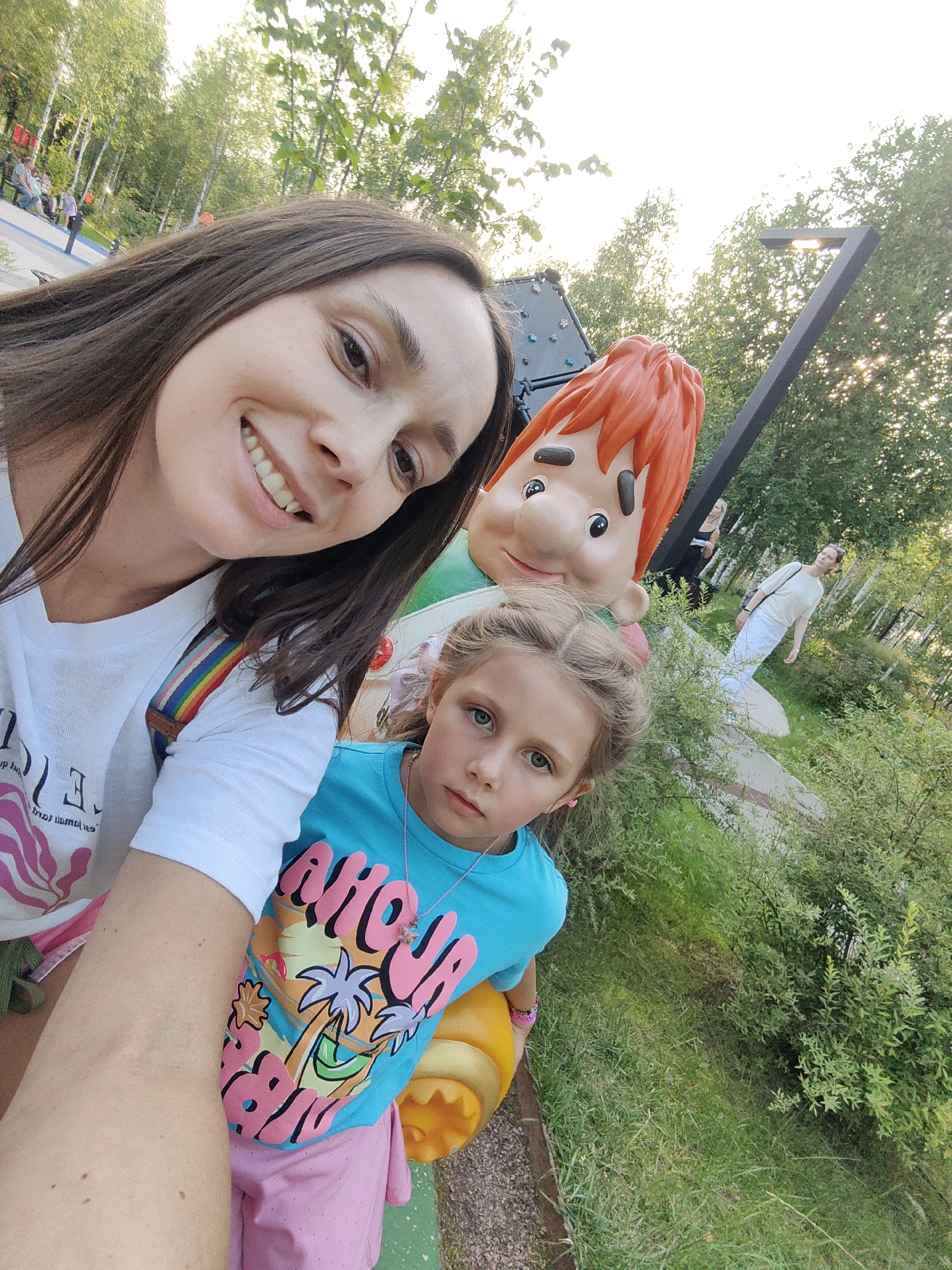 A selfie captures a woman and a young girl in an outdoor park setting. The woman, with long brown hair, smiles broadly at the camera, wearing a white t-shirt with a rainbow-striped strap visible. Beside her, the young girl, with blonde hair pulled back, looks towards the camera with a neutral expression, wearing a blue t-shirt with a tropical graphic and a necklace. Behind them, a large cartoon character figure with orange hair stands. In the background, trees and lush green bushes are visible, along with a modern-looking light fixture. Further in the distance, two women are seen walking. The scene appears to be a family-friendly outdoor space, possibly a children's play area or park. This photograph was taken in Gorky Park in Moscow, Russia.