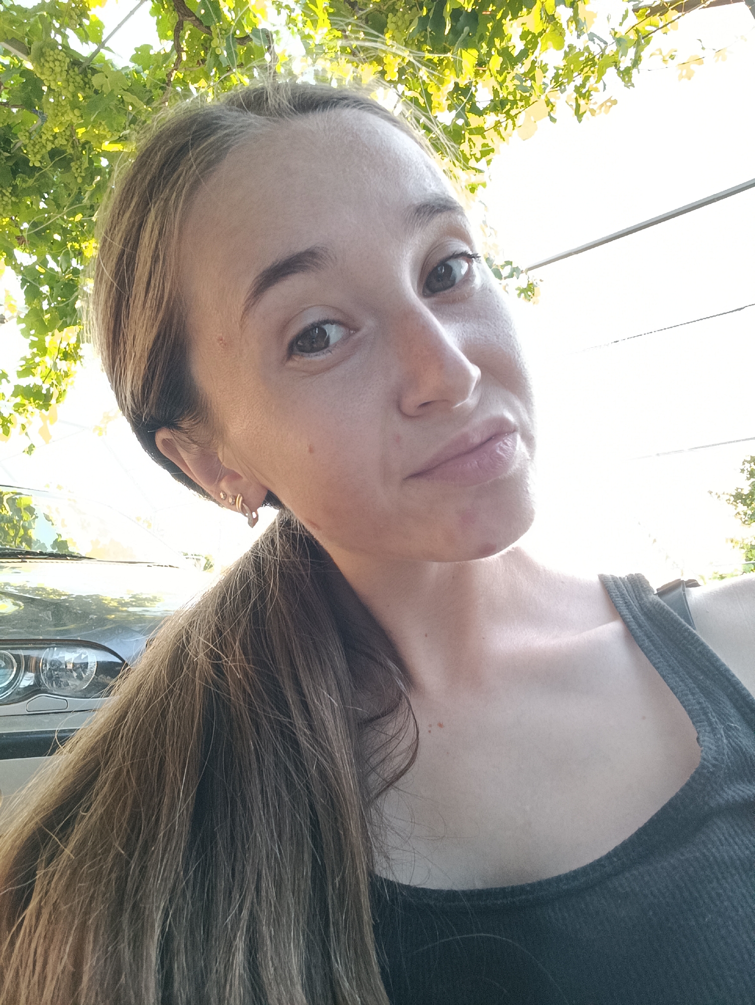 A woman is captured in a close-up, outdoor selfie. She is looking towards the camera with a slightly pursed-lip expression. Her hair is long and light brown, pulled back and falling over her shoulder. She is wearing a dark-colored tank top. In the background, green foliage and grapes are visible overhead, suggesting a vineyard or garden setting. Part of a car is visible on the left side of the frame, with its headlight and grille showing. The lighting indicates it is daytime, with the sun filtering through the leaves, creating a bright background.