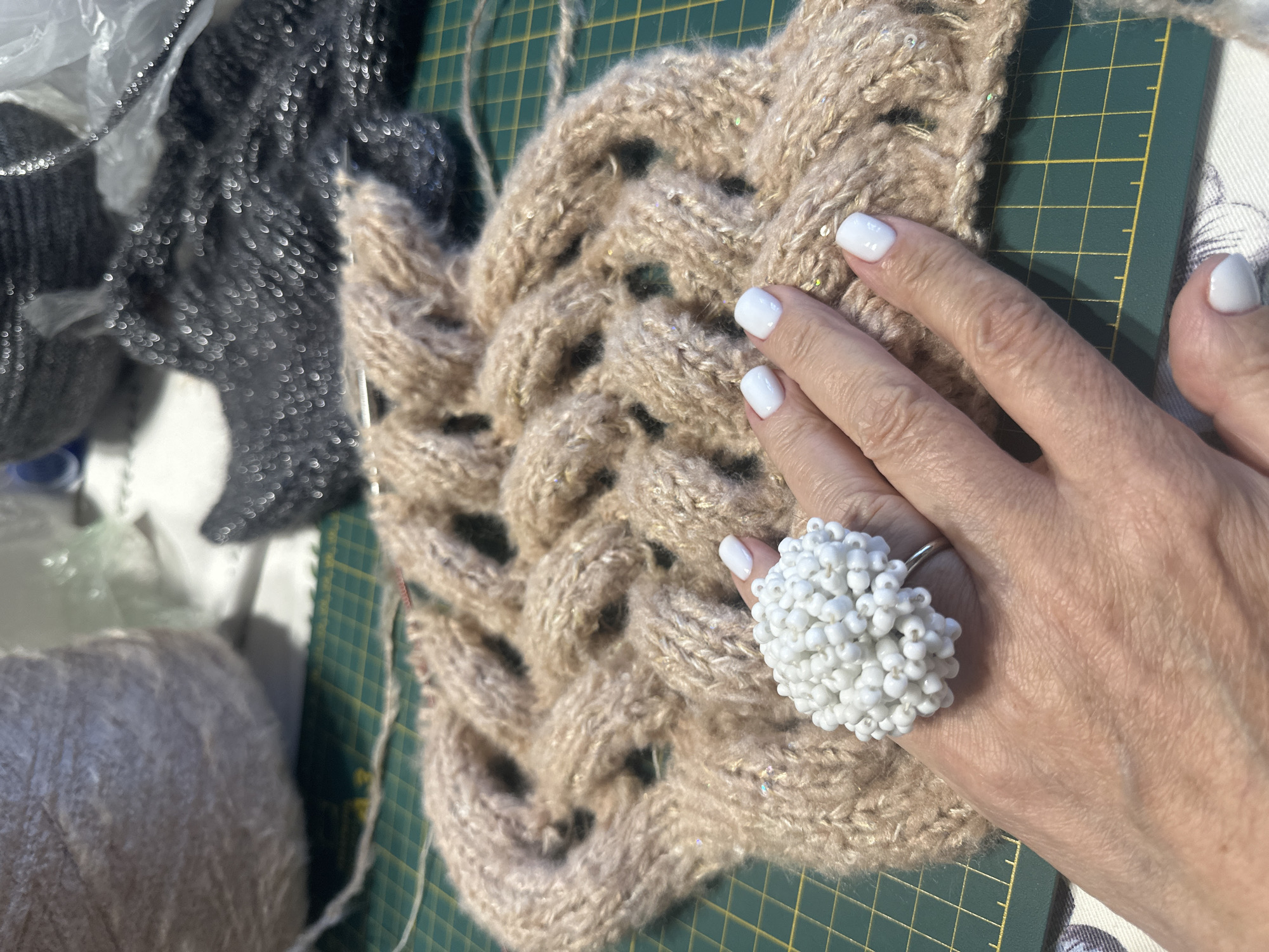 An overhead shot shows a hand with white nail polish and a large white beaded ring resting on a piece of light brown cable-knit fabric with gold thread accents. The fabric is on a green cutting mat with yellow grid lines. In the background, there are balls of yarn and dark gray sparkly fabric.