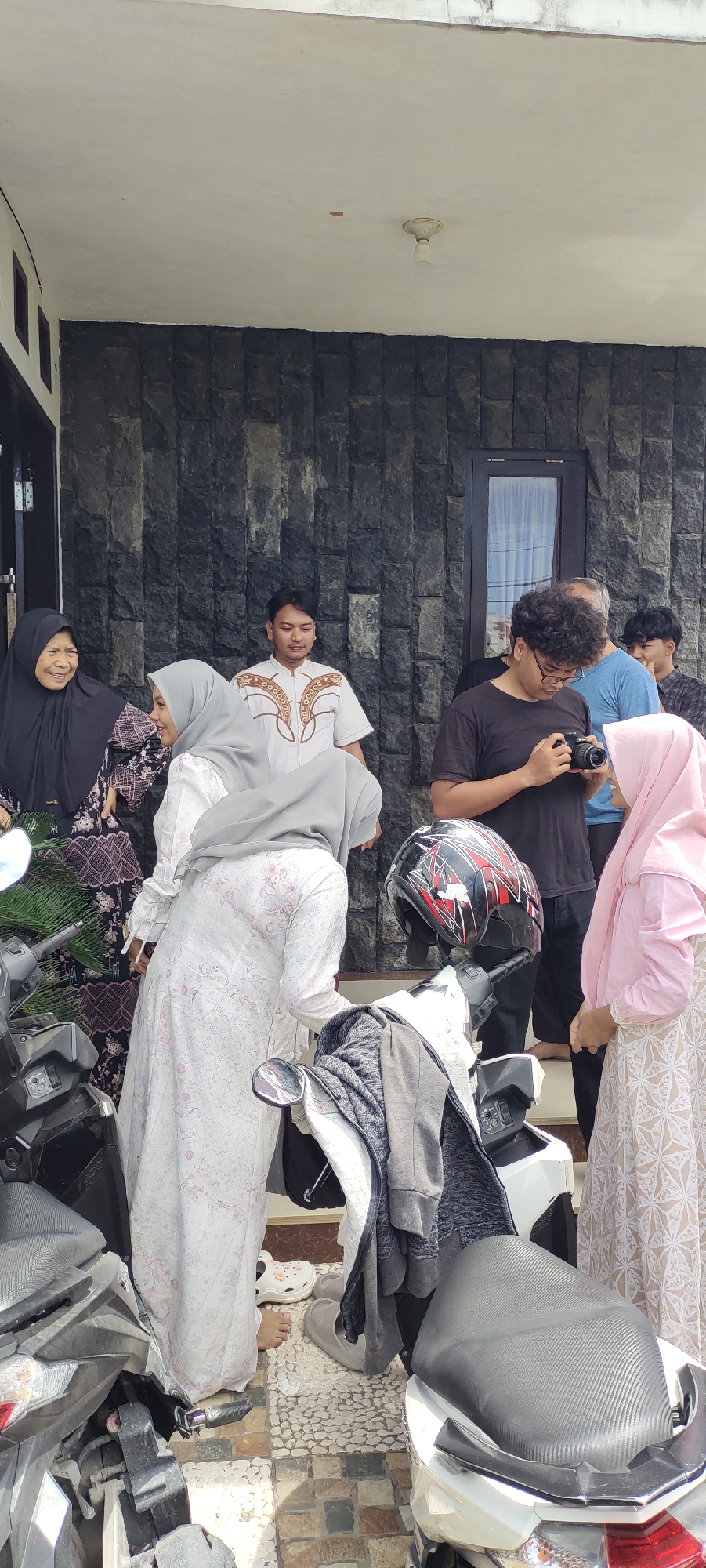 The image shows a group of approximately 8 people gathered outside a building with dark stone cladding in Lhokseumawe, Indonesia. The scene appears to be informal, possibly a gathering of family or friends. Several individuals are wearing Muslim headscarves (hijabs). One person is holding a camera and seems to be taking pictures. A parked motorcycle with a helmet resting on it is visible in the foreground, partially obscuring the view. The setting appears to be a residential area. The ground is tiled.