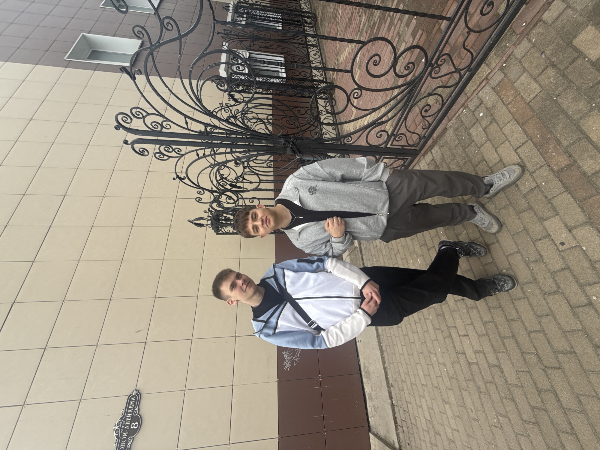 Two young men stand next to each other. The man in front is wearing a white, black, and light blue jacket with black pants. The man behind him is wearing a gray jacket, black shirt, and gray pants. They are standing on a brick sidewalk in front of a light tan building with dark brown trim. A black wrought iron gate is behind them. A street sign reads "ул. Алейхема, 8" which translates to 8 Sholem Aleichem Street, Birobidzhan, Russia.