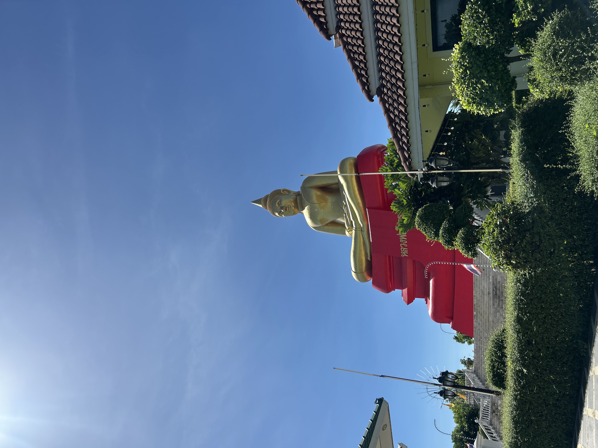 A large, gold Buddha statue sits atop a bright red, tiered platform in Thai Ko Subdistrict, Thailand. The Buddha is in a seated meditation pose with its legs crossed. The statue is golden and shiny, reflecting the bright sunlight. The red platform has some Thai script on its front. To the right of the Buddha statue is a building with a brown tiled roof. In front of the building and the Buddha are manicured green bushes and trees. A set of stone stairs leads up to the Buddha statue from the foreground. The sky is a clear, bright blue with no clouds visible. The overall scene depicts a religious monument on a sunny day in Thailand.