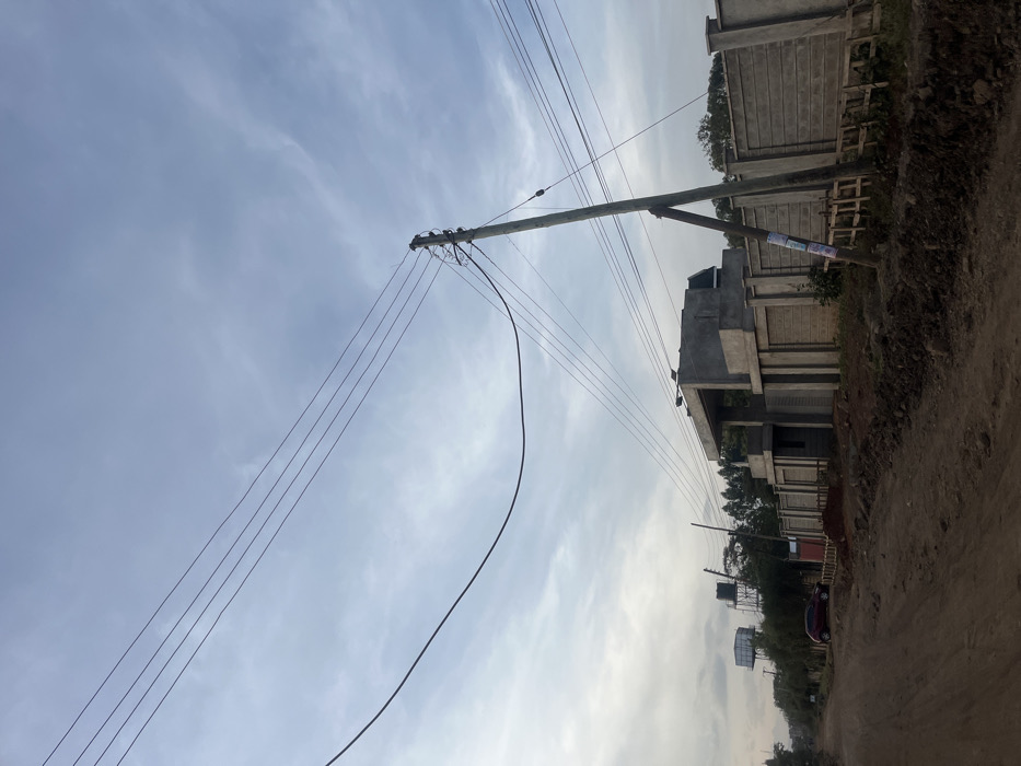 The image displays an outdoor scene in Gitothua ward, Kenya, under a cloudy sky. An unpaved dirt road is visible in the foreground and extends into the distance, bordered by dark soil piles. A prominent wooden utility pole with a diagonal wooden brace stands on the right side of the road, supporting numerous electrical power lines that crisscross the upper portion of the frame. To the right of the utility pole, a large, unpainted concrete structure, consistent with a building under construction, is situated behind a perimeter fence constructed from light grey precast concrete panels. Wooden planks are propped against this fence near the pole. Further along the dirt road to the left, a dark red vehicle, resembling a hatchback, is parked. In the background on the left, two elevated water tanks on metal support structures are visible above a tree line. No individuals are present in the scene.
