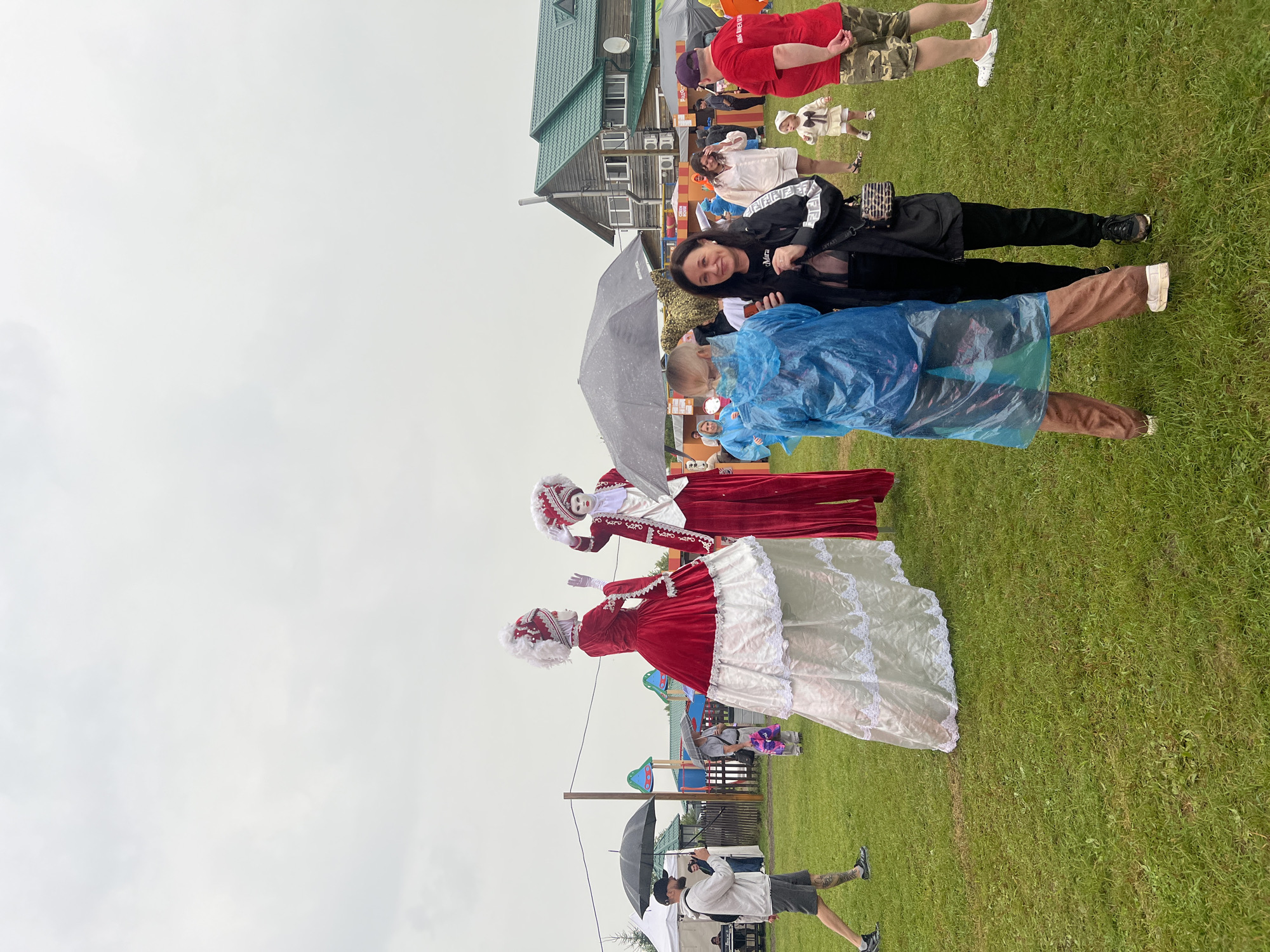 In this outdoor scene, several people are gathered on a grassy area, possibly during an event. Two large figures in elaborate red and white historical costumes stand prominently in the foreground. One figure, to the left, is dressed in a long red gown with white lace trim and a white wig, holding their hands up. The other figure, to the right, wears a similar red and white outfit, with a white wig and white gloves.
In the mid-ground, a woman in a black jacket with white lettering and a crossbody bag is looking at the camera with a smile, holding an umbrella. Beside her, a child wearing a blue poncho is also visible, holding an umbrella. Further in the background, more people are scattered, some holding umbrellas, suggesting it might be raining or overcast. A wooden building with a green roof is visible to the left, and in the distance, playground equipment can be seen.
The overall mood appears festive, despite the possibly inclement weather. The presence of costumed figures and umbrellas suggests a public gathering or festival. The lighting indicates it is daytime. Some visible text on the woman's jacket reads "FENDI". The location is indicated as Кузнецово, Russia.