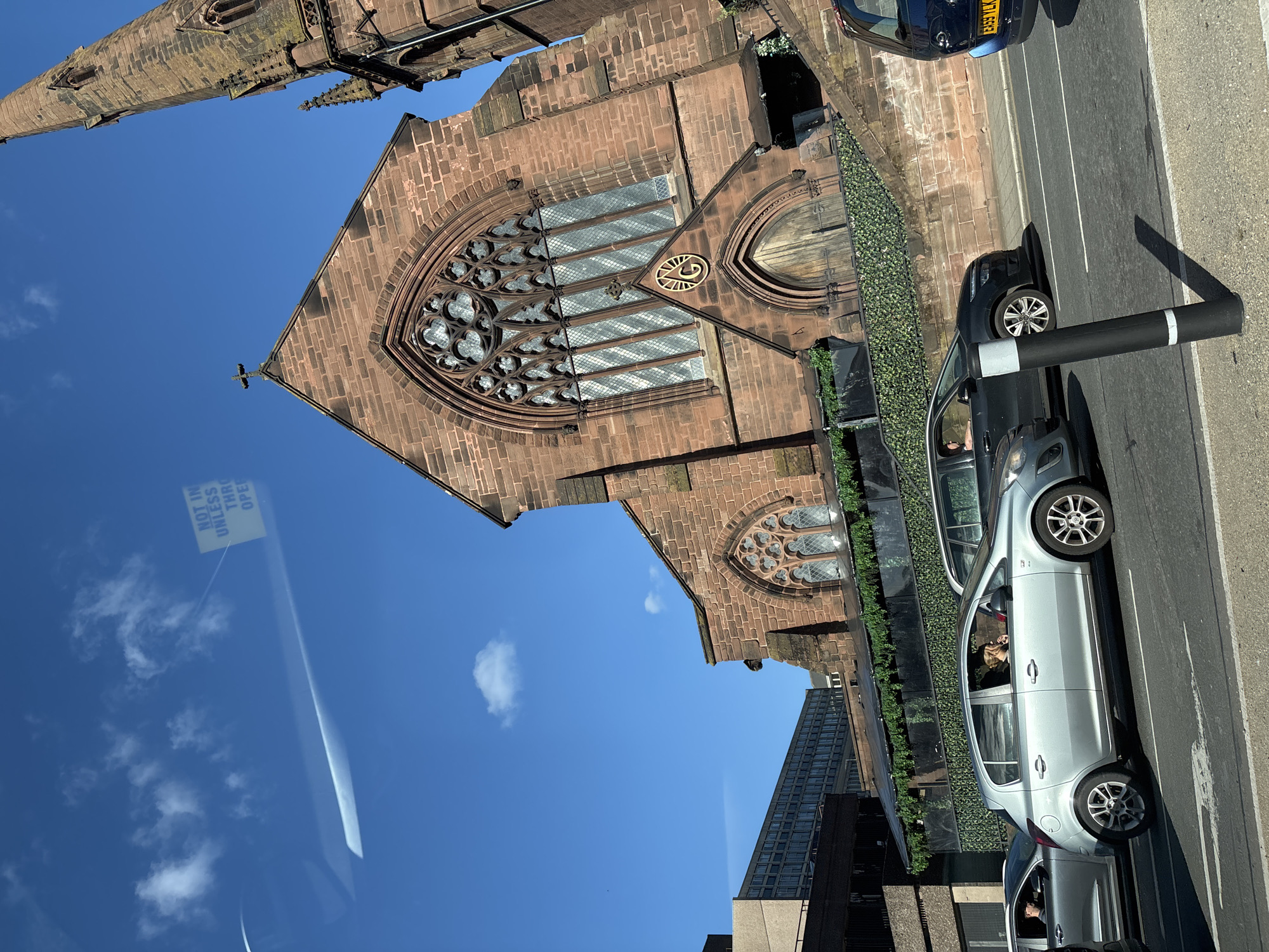 The image captures a view of the ruins of the old Coventry Cathedral in Chester, United Kingdom, taken from inside a vehicle. The cathedral's partial spire and the main section of its facade with a large, ornate window are visible against a clear blue sky with some cloud cover.
In the foreground, there are several cars parked alongside the road. One silver car appears to be occupied, and a black bollard stands beside the road. The scene is in an urban environment, with parts of modern buildings visible in the background. The image suggests a blend of historical ruins juxtaposed with contemporary urban life.
