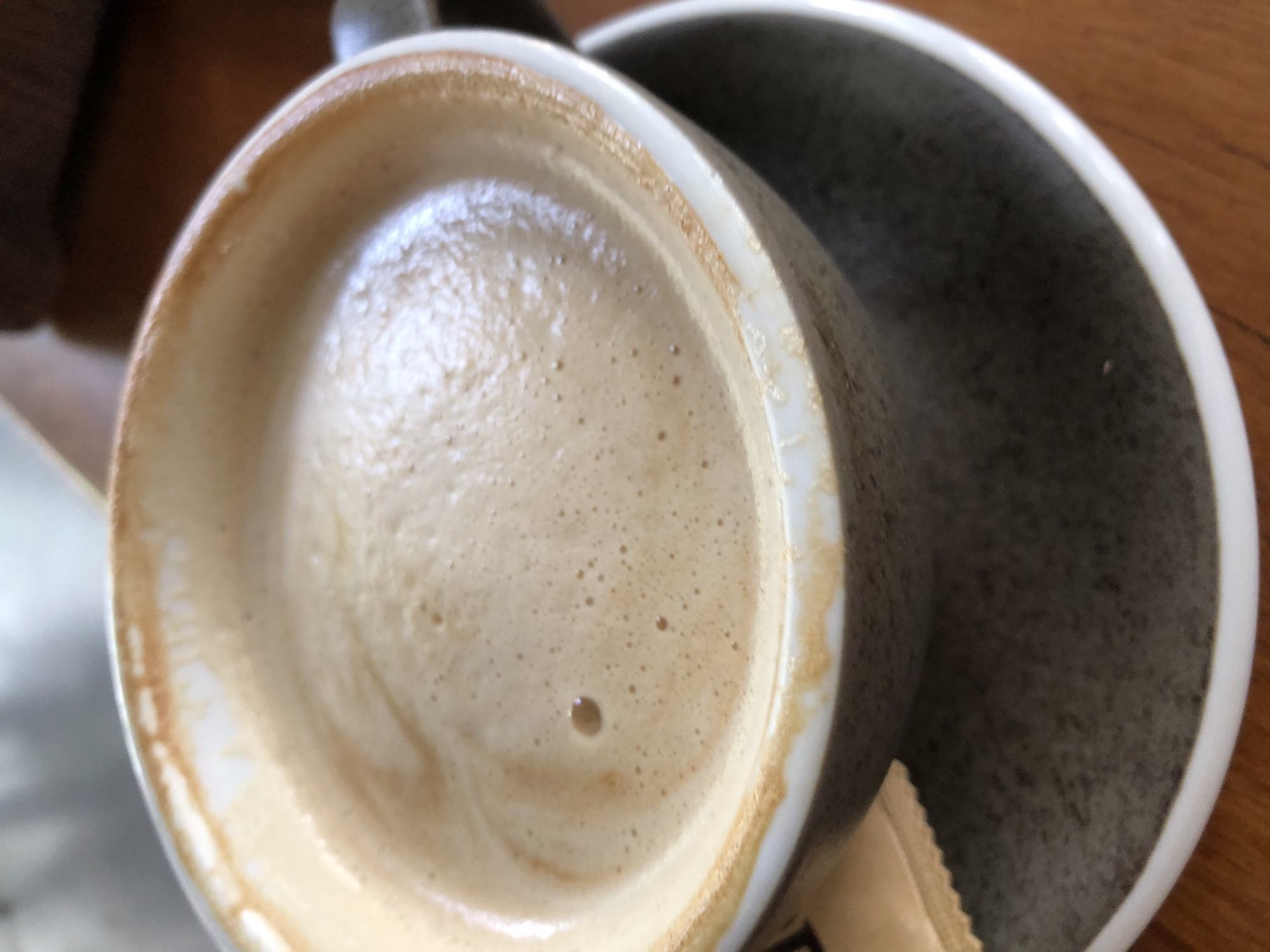 A close-up, eye-level shot captures a cup of coffee with foamy milk, resting on a saucer. The coffee is a light brown color with a frothy top, showing a few small bubbles and a single larger bubble in the center. The rim of the white ceramic cup has a slightly worn, brownish edge. A small, beige sugar packet is partially visible to the left of the cup, peeking out from under its rim. The saucer, also ceramic, has a speckled grey pattern with a white lip. The coffee cup and saucer are placed on a wooden surface, likely a table. The background is blurred, suggesting an indoor setting, possibly a cafe. There are no people visible in the image. The overall impression is one of a simple, unadorned moment of enjoying a beverage. This scene could be found in many cafes across the vibrant city of Đà Nẵng, Vietnam, where coffee culture is prevalent and a cup of coffee is a common sight on café tables.