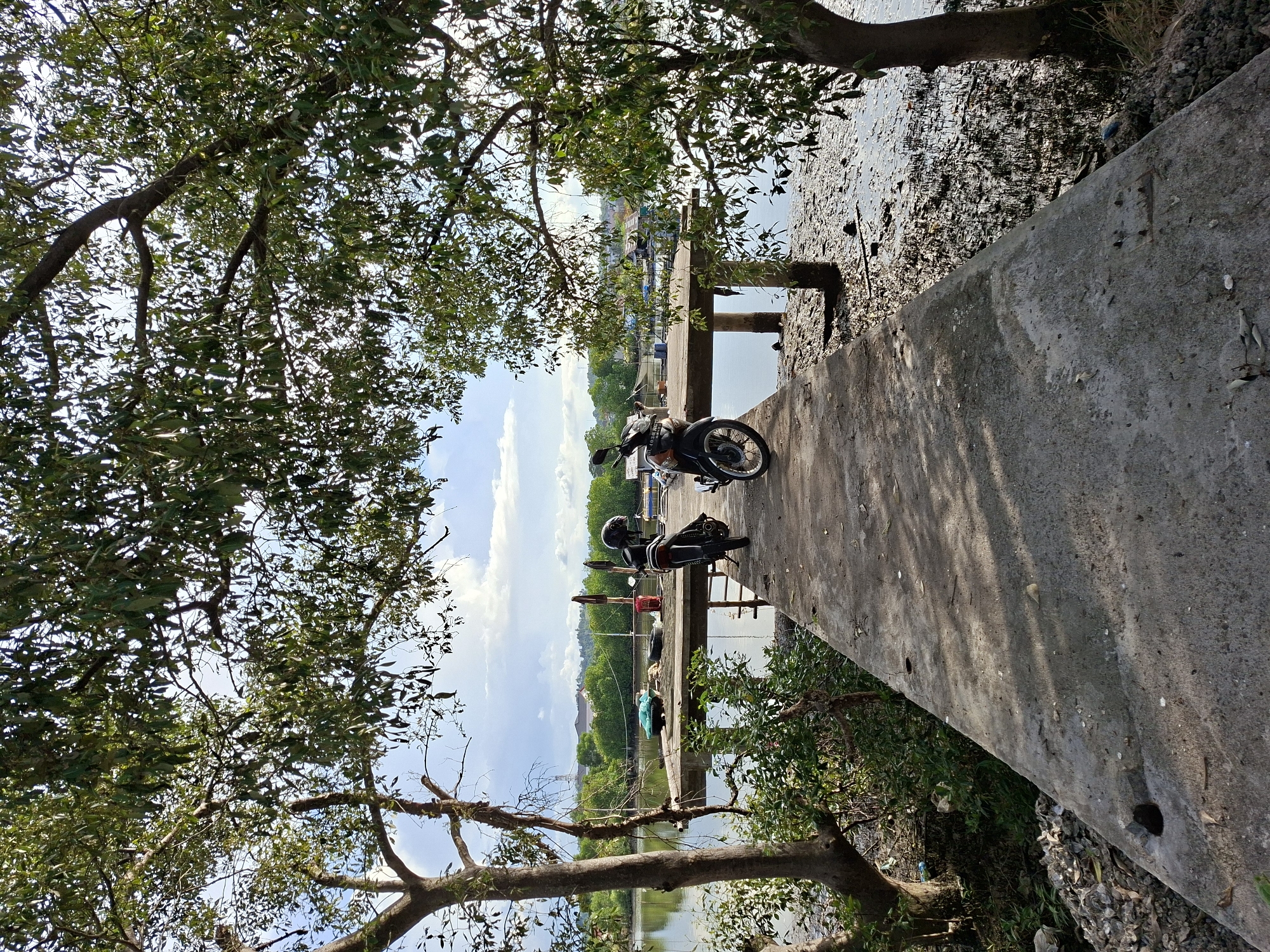 The image shows a motorcycle parked on a concrete pier in Lhokseumawe, Indonesia. The pier extends into the water, with a muddy shoreline visible on one side. Trees with dense foliage frame the scene. The sky is partly cloudy, with blue patches visible. The photo appears to be taken during the day. A helmet rests on the motorcycle. In the distance, there are more buildings and structures, presumably within Lhokseumawe.