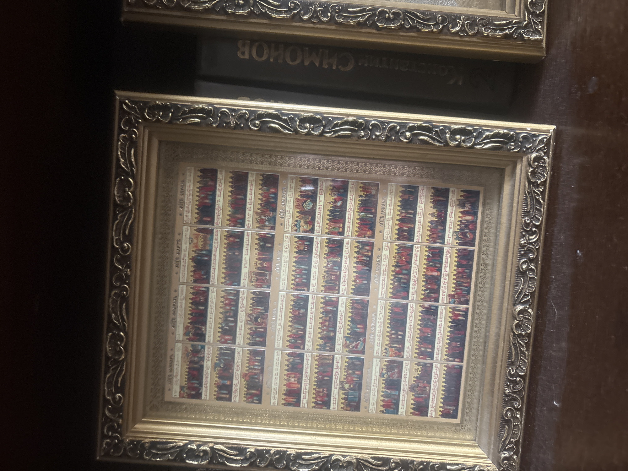 A framed collection of small religious icons, likely depicting saints or biblical scenes, is displayed on a wooden surface. The frame is ornate and golden with decorative carvings. To the right of the framed icons, a dark-bound book with gold lettering is partially visible. The book appears to be in Cyrillic script, with the visible text reading "КОНСТАНТИН СИМОНОВ". The overall scene suggests an indoor setting, possibly a home or a religious establishment, with the items arranged on a shelf or table. The photograph is taken from a slightly elevated angle, focusing on the framed artwork and the adjacent book. The background is dark and out of focus, making it difficult to discern specific environmental details. No people are visible in the frame.