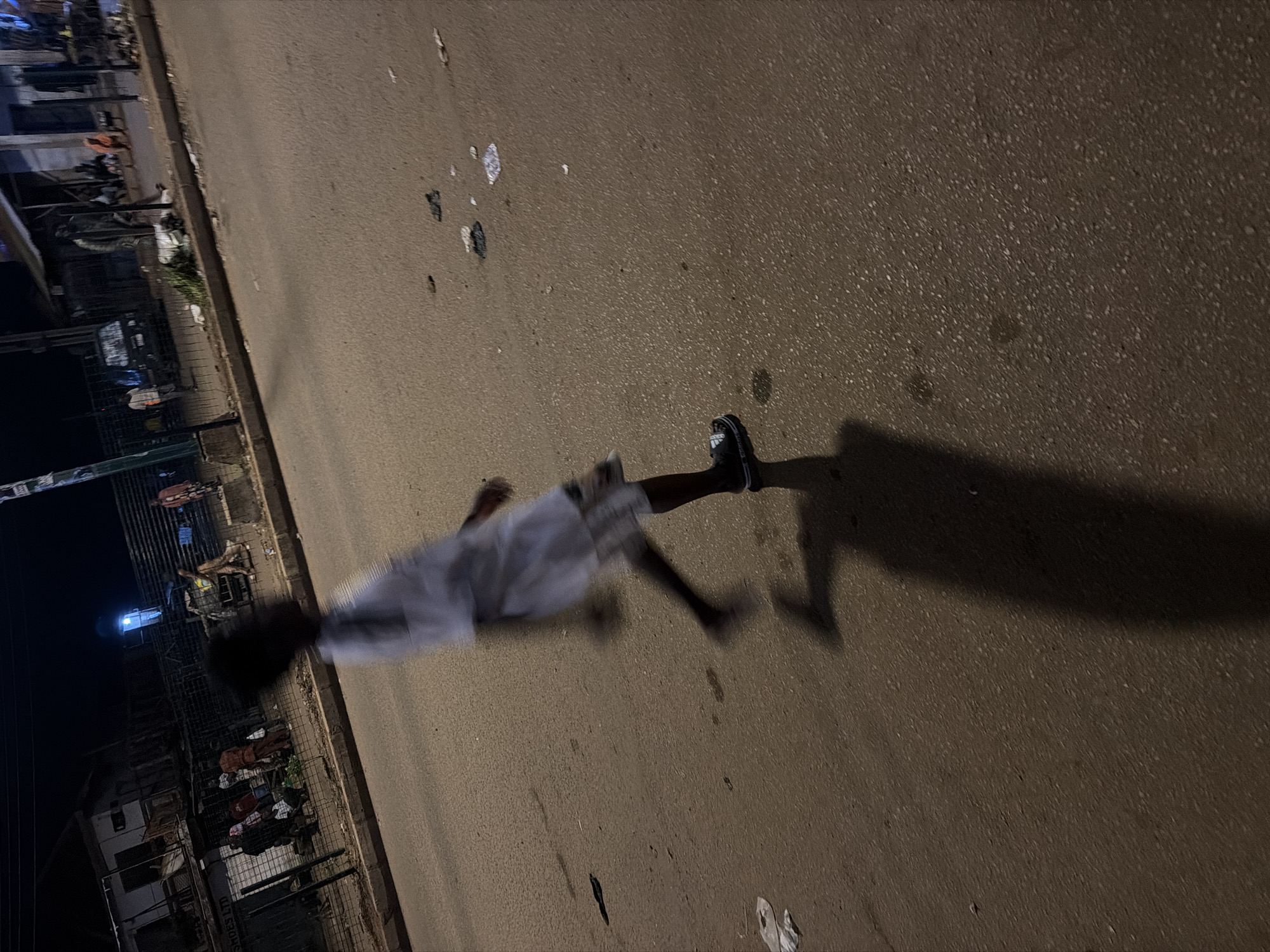 The image depicts a blurry night scene on a paved street in Bukuru, Nigeria. A young person wearing a white shirt and patterned shorts is captured mid-stride, with their motion creating a blurred effect. Their shadow is cast long on the asphalt. In the background, several buildings line the street, some appearing to be shops or residences. A few individuals are visible near the buildings, suggesting activity on the sidewalk. The street itself is littered with debris, including pieces of paper and other small objects. A parked vehicle is also present in the background. The lighting appears to be from streetlights and the ambient light from the businesses.