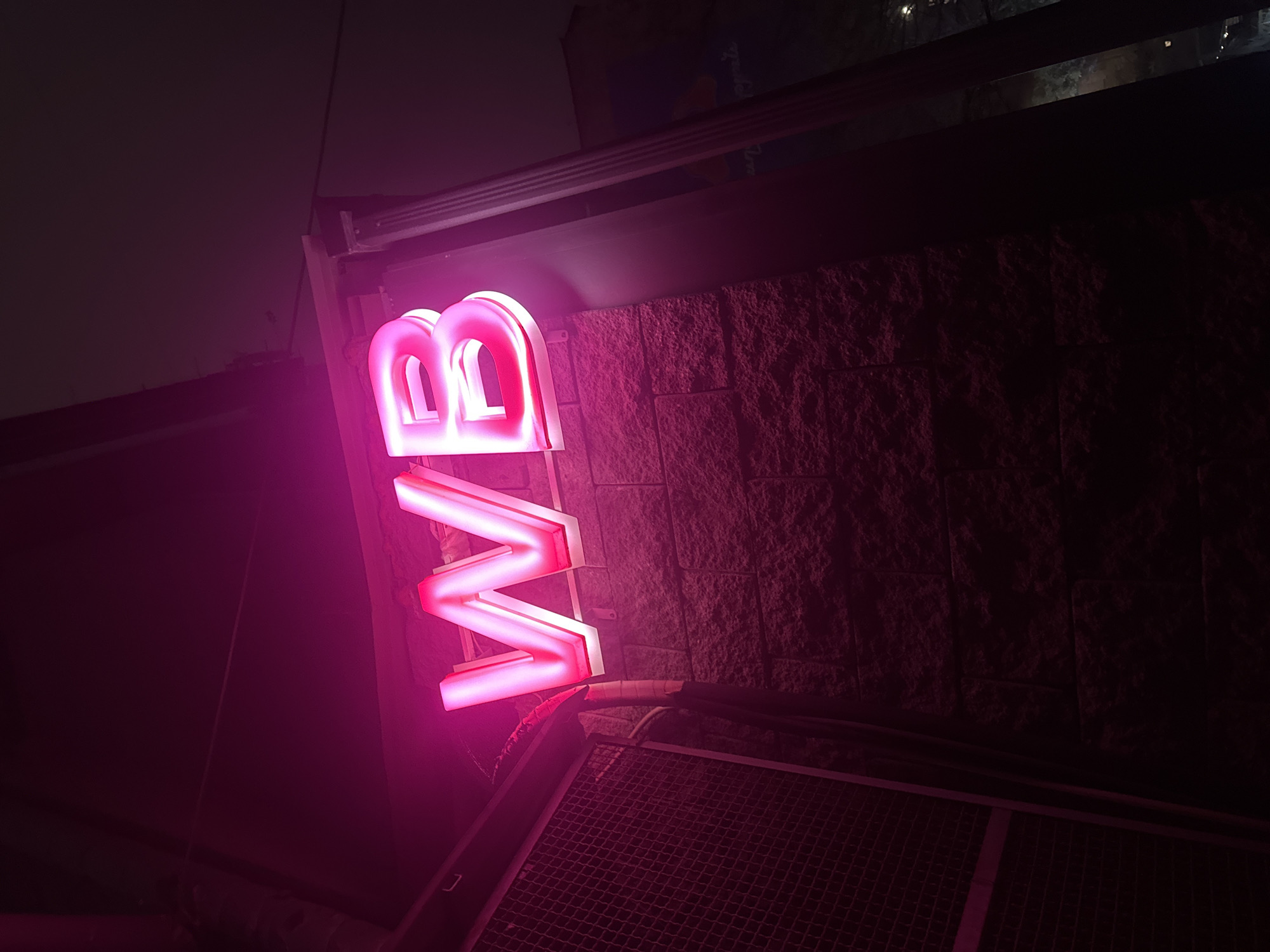Captured at night in an unidentified city, the image focuses on a brightly lit pink neon sign reading "WB". The sign is affixed to a wall constructed of rough, dark stone blocks. Below the sign, a metal grid or grate is visible, its dark surface reflecting the sign's pink glow. The surrounding environment is dark, with only faint hints of cityscape or other structures in the background. The photograph appears to be taken from a low angle, looking upwards towards the sign.