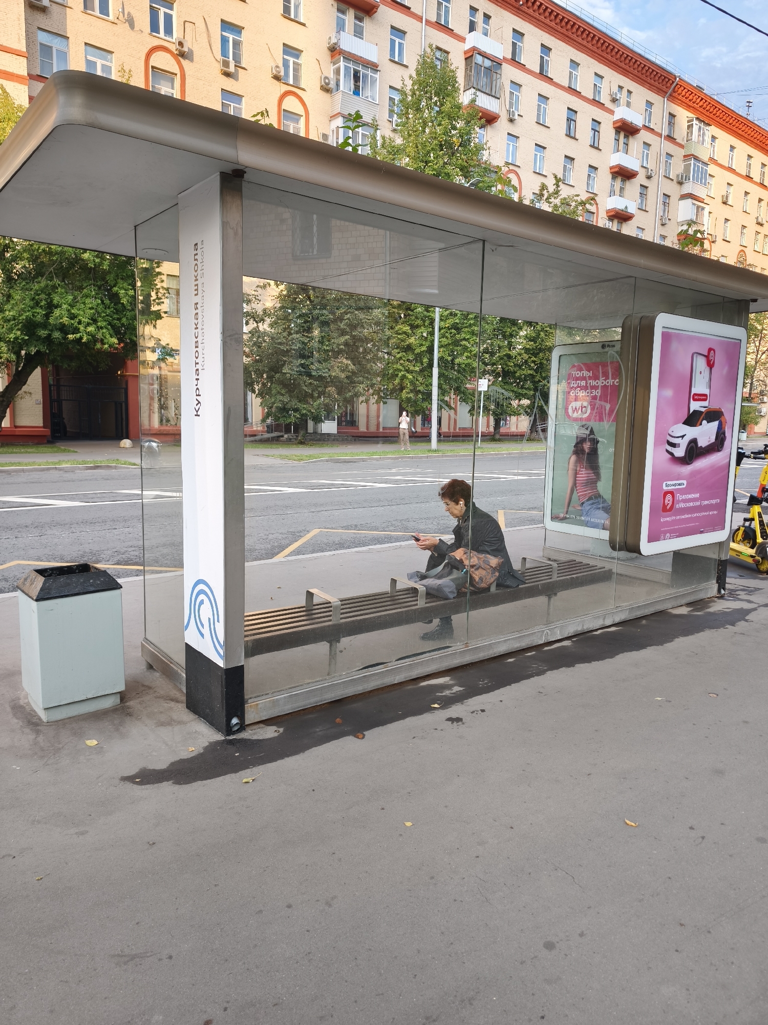 This is a medium shot of a bus stop shelter in Moscow, Russia. A woman is sitting on the bench inside the shelter, looking at her phone. She is wearing a black coat and has short brown hair. There is a trash can next to the bus stop. A yellow electric scooter is parked to the right of the bus stop. The bus stop has glass walls and a metal roof. An advertisement for a transportation app is displayed on the right side of the bus stop. The background shows a building with red and beige facades, trees, and a street. The sky is overcast.
