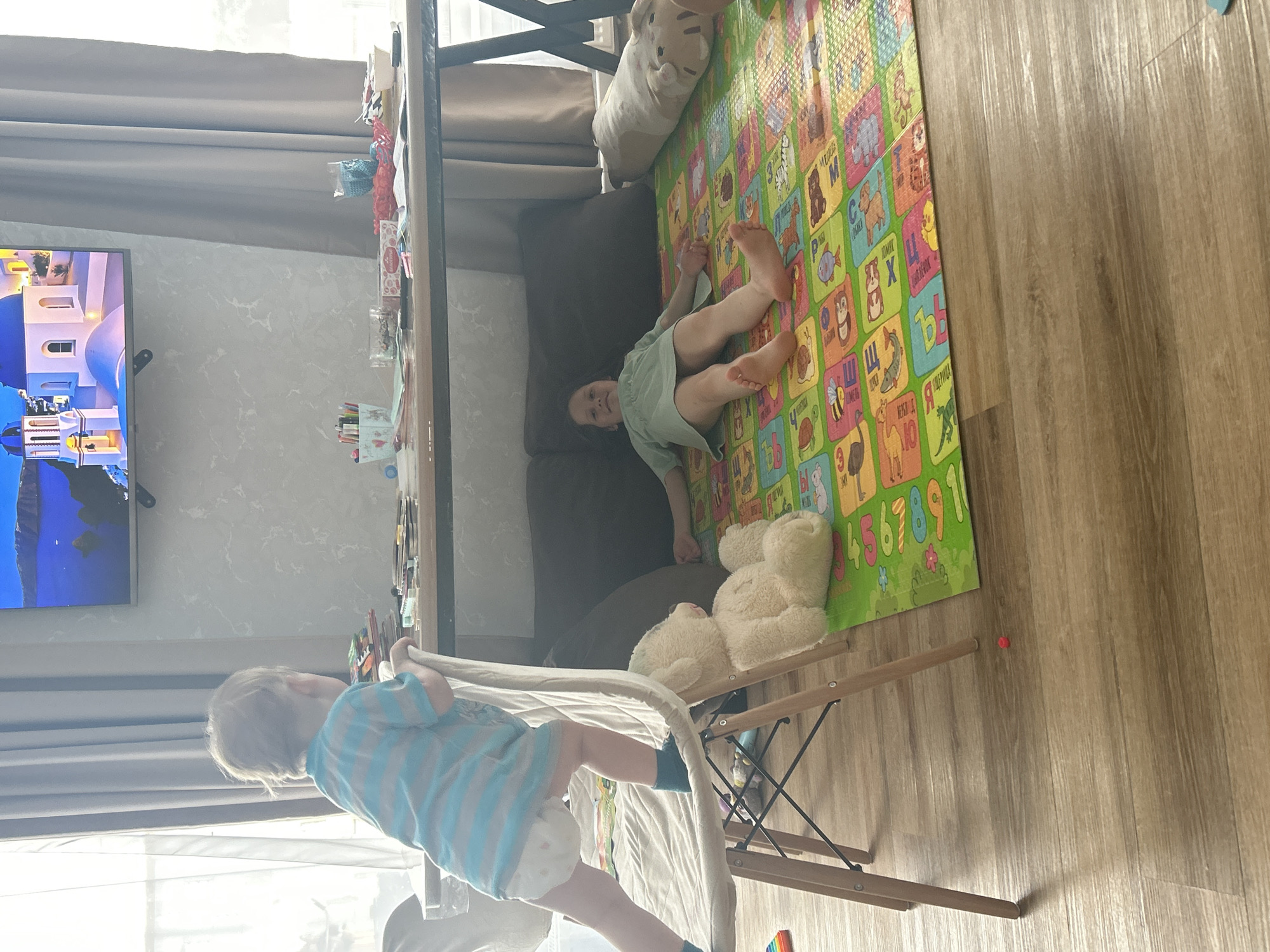 The image shows an interior scene in Chaikovsky, Russia. A young child in a blue and white striped shirt and diaper is standing near a folding wooden drying rack covered with a white sheet. Another child is lying on a colorful alphabet play mat on the floor. A brown sofa and a table with objects on it are also visible. A TV screen on the wall displays a scene of Santorini, Greece. A stuffed animal is on the mat, and the floor is light brown wood.