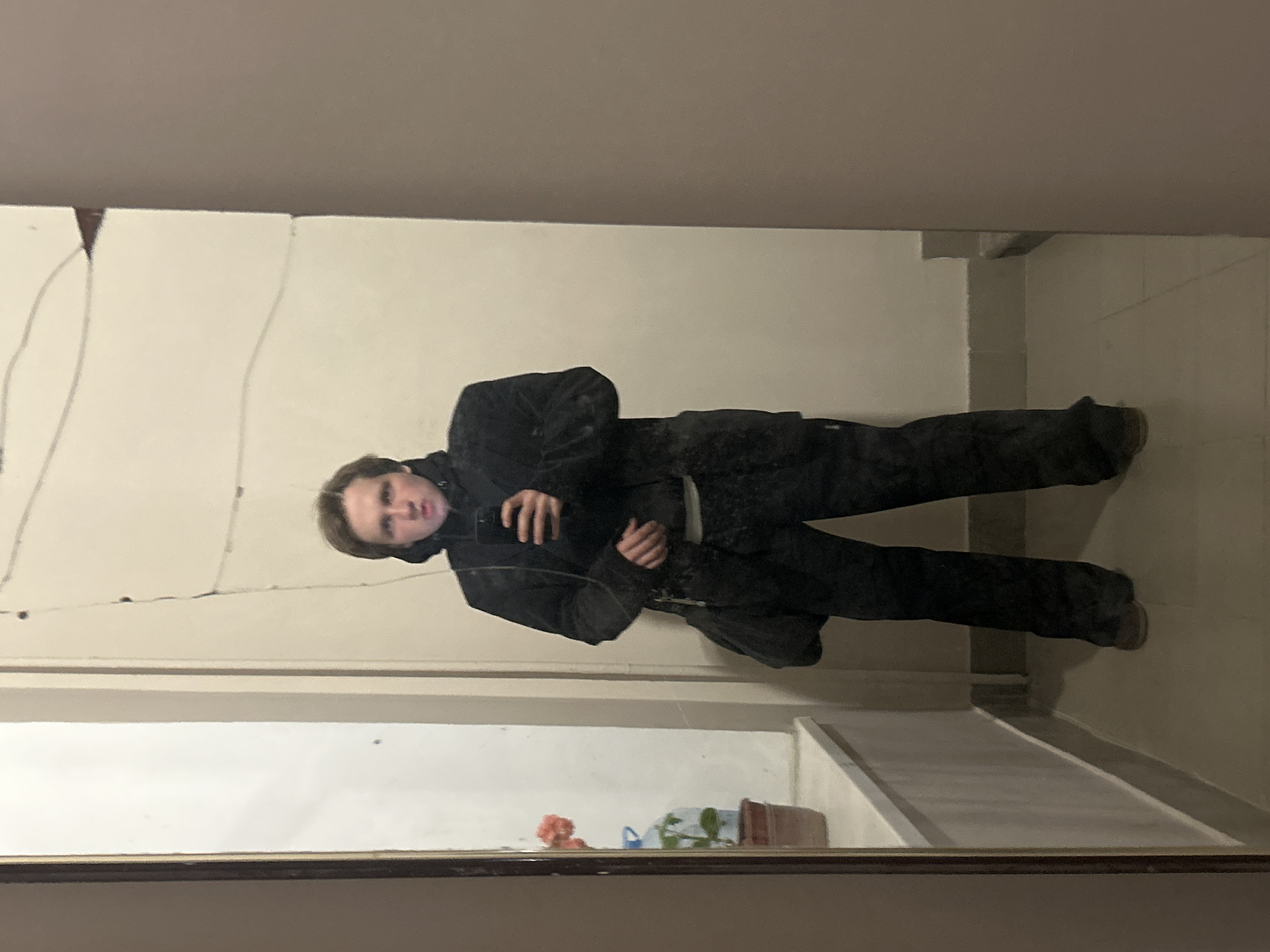 A person in dark clothing stands in front of a cracked mirror, holding a cell phone. The background reveals an interior space with light-colored walls. On a shelf below the mirror, there's a flower arrangement and a toppled plant pot. The city and specific location are unknown.