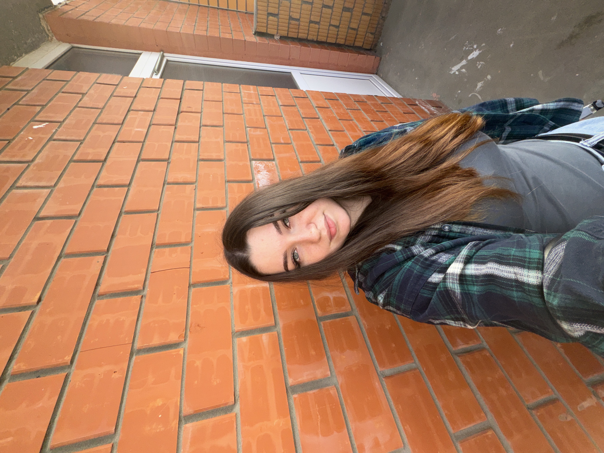 A young woman with long, brown hair takes a selfie outdoors, leaning against an orange brick wall. She is wearing a grey t-shirt and a dark green and blue plaid shirt. Her hair is long and falls over her shoulder and down her back. She is smiling slightly and looking towards the camera. The background consists of the brick wall, a portion of a window with white trim, and a concrete surface above. The lighting suggests it could be daytime, possibly overcast given the lack of harsh shadows. The scene appears to be an outdoor, urban or semi-urban environment. No text is visible in the image.