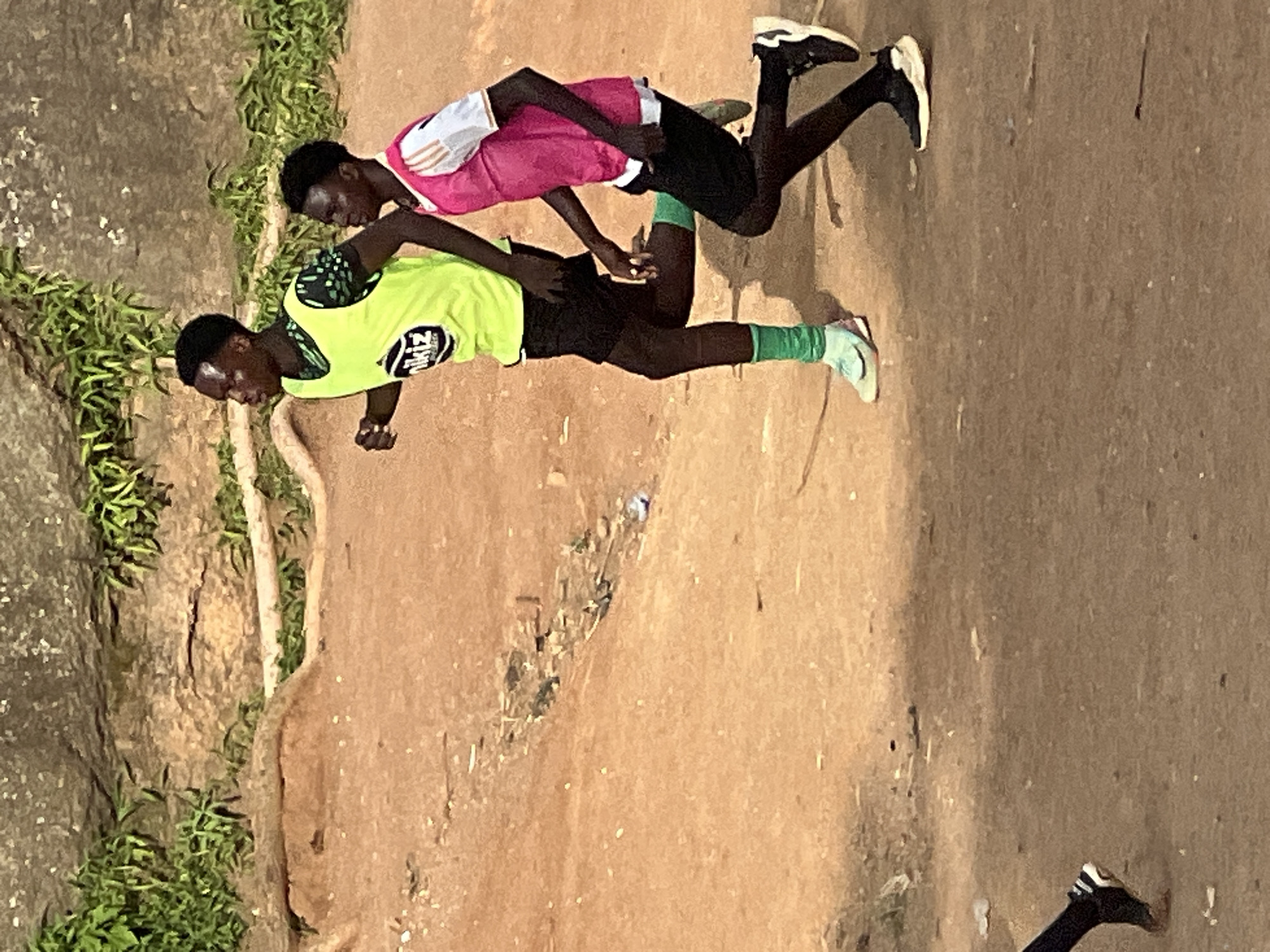 Two young Black males are running side-by-side on a dirt path. The male on the left is wearing a bright green vest over a black t-shirt with a green and black pattern, black shorts, and light blue athletic shoes with pink accents. He has green socks that go up to his calves. The male on the right is wearing a pink vest over a white t-shirt with orange trim, black shorts, and black and white athletic shoes. Both individuals appear to be engaged in physical training. The background consists of a rocky, uneven hillside with sparse green vegetation. The ground is dry and dusty, with some scattered debris. This scene is consistent with an outdoor training ground in a more natural or less developed area, which could be found on the outskirts of a city like Jos, Nigeria, where open spaces and rugged terrain are common.