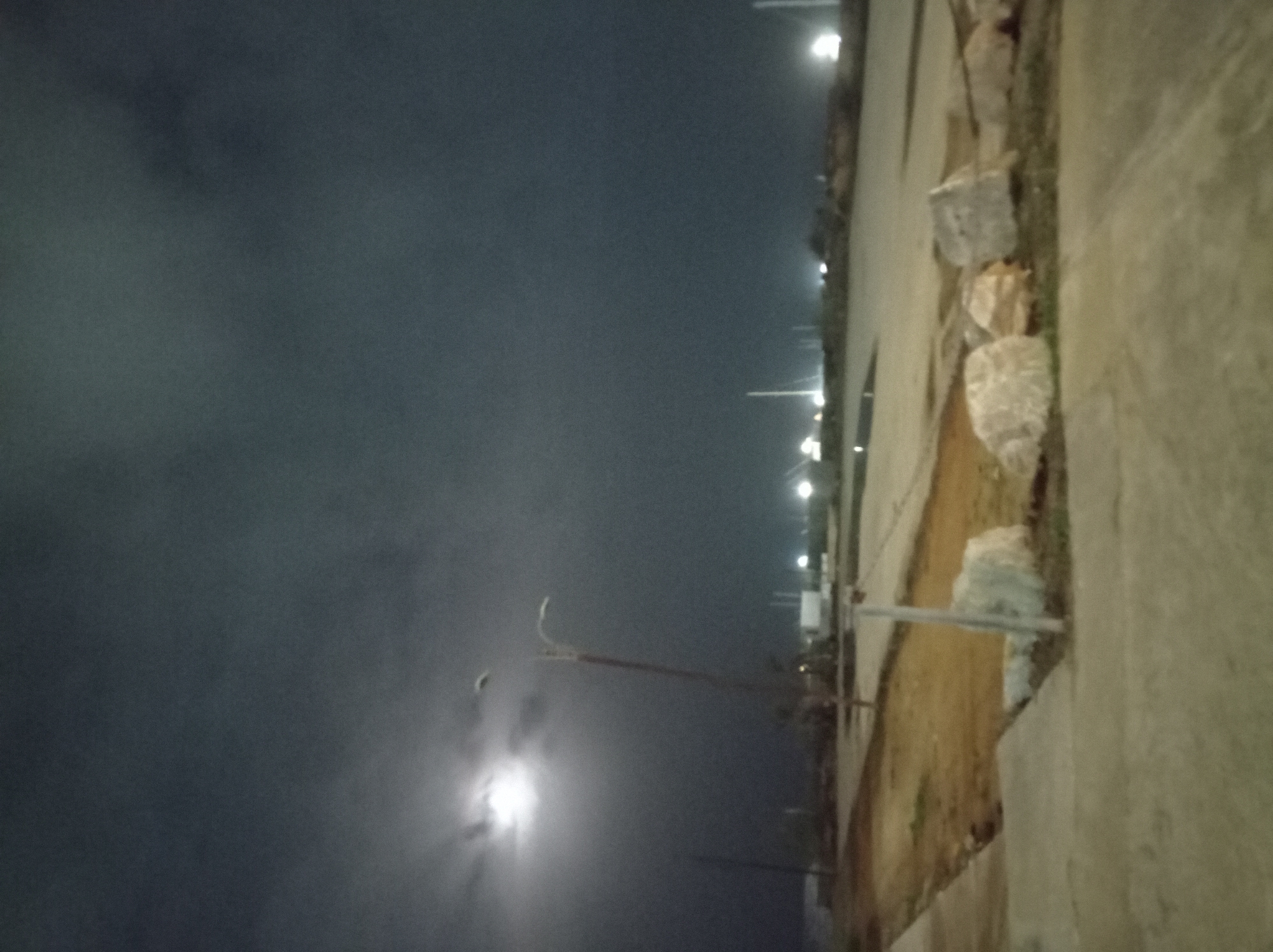 The image displays a nighttime outdoor scene, likely along a road or pathway, under a dark and partially clouded sky.
In the upper-left portion of the frame, a bright, luminous moon is prominently visible, its light diffused by atmospheric haze or thin clouds, creating a distinct halo effect. The sky is a deep, dark grey-blue, indicating the absence of daylight, with subtle variations suggesting cloud cover.
A tall, slender pole, likely a lamppost, stands in the mid-left area, extending upwards into the dark sky. Its top curves, suggesting a fixture for a light source, though the light itself is not directly captured.
Across the mid-right of the image, a series of bright, distant artificial lights punctuate the darkness, appearing as glowing points. These lights are aligned, hinting at a distant street, road, or developed area.
In the foreground, running along the bottom and right edge of the image, is a paved surface, likely a concrete path or sidewalk. Adjacent to this path, and acting as a divider from the immediate surroundings, are several large, light-colored rocks or chunks of debris. Beyond these rocks, to the left of the path, lies an area of reddish-brown earth or dirt, suggesting an unpaved shoulder, a vacant lot, or an area under development.
The scene is devoid of people or any specific activity, presenting a quiet, static nocturnal view. No visible text can be discerned. The combination of artificial streetlights, the natural moonlight, and the mix of paved and unpaved terrain suggests a semi-urban or transitional environment, characteristic of an area like Porlamar, Venezuela.