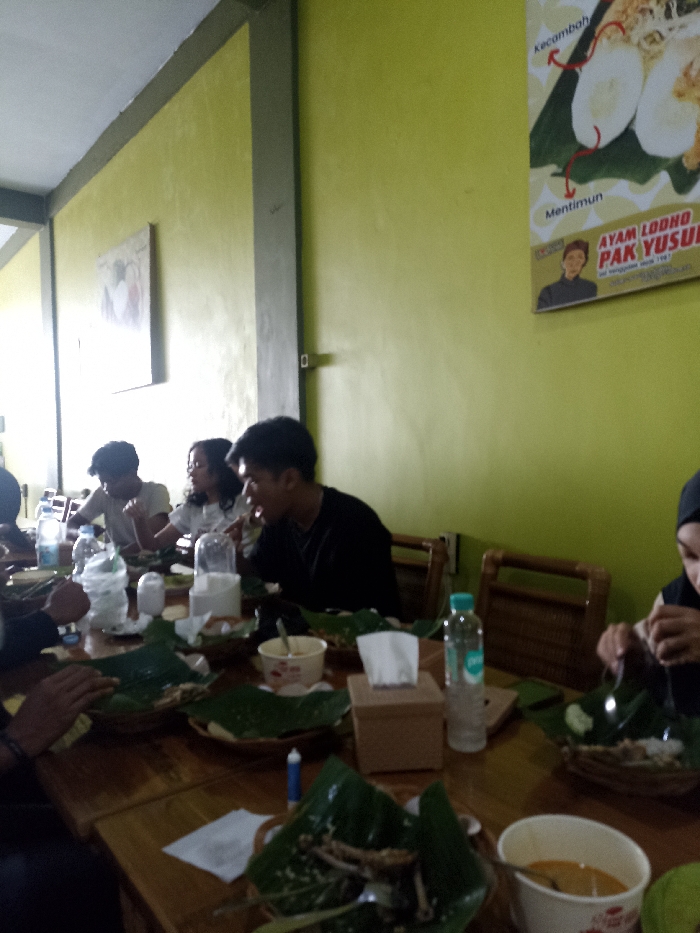This image captures a group of individuals enjoying a meal together at an indoor restaurant or eatery in Tulungagung, Indonesia. The scene is set around a large wooden table, surrounded by bamboo-style chairs. The walls of the establishment are painted a vibrant yellow or lime green.
There are at least six people visible, some more clearly than others. They appear to be young adults or a mix of ages, dressed casually. The central activity revolves around eating and socializing. Each person has a meal served on a banana leaf placed within a woven basket, a traditional Indonesian presentation style. Various food items, possibly chicken and rice, are visible on these leaves, along with some side dishes in small bowls. Bottled water is prominent on the table, along with a tissue box, indicating a casual dining setting. Some individuals are actively eating, with expressions suggesting contentment and focus on their food, while others appear to be conversing.
Notable details include the bright lighting, suggesting it's daytime. The overall atmosphere appears relaxed and communal. On the wall in the upper right, there's a poster advertising a dish. Visible text on the poster includes "AYAM LODHO PAK YUSUF," which translates to "Pak Yusuf's Lodho Chicken," suggesting the restaurant specializes in this Javanese dish. Below the main dish illustration, smaller labels point to ingredients like "kecambah" (bean sprouts) and "Mentimun" (cucumber). Another framed picture hangs on the wall further to the left. The serving style and the advertised dish strongly indicate a local Indonesian culinary experience.