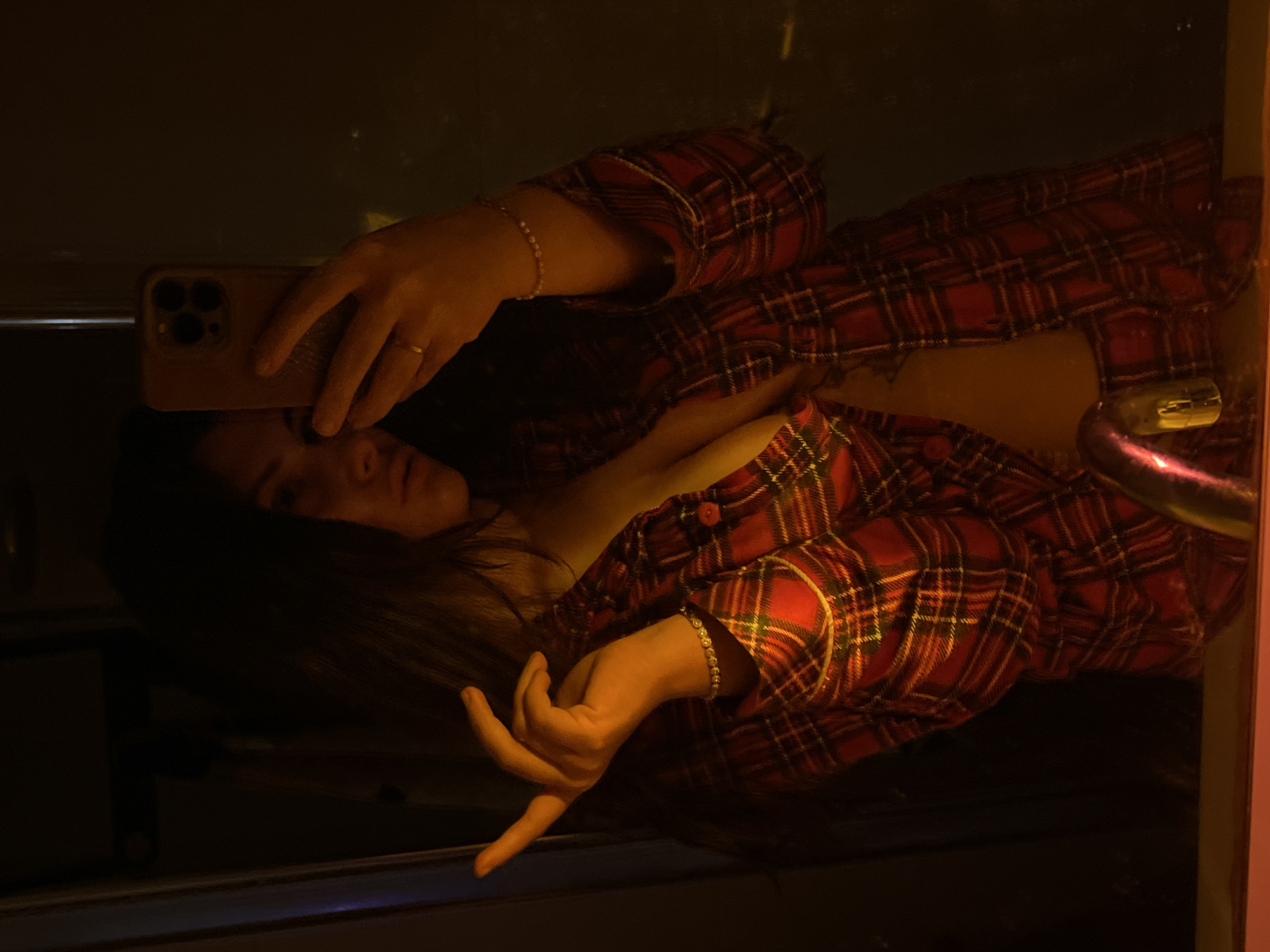 A woman is taking a selfie in a mirror. She is wearing a red and black plaid shirt, unbuttoned. She is holding a cell phone with her right hand. She is wearing a ring on her right hand. Her hair is dark and long. The setting appears to be indoors. There is no verifiable information in the image to support a connection to Kiryat Bialik, Israel.