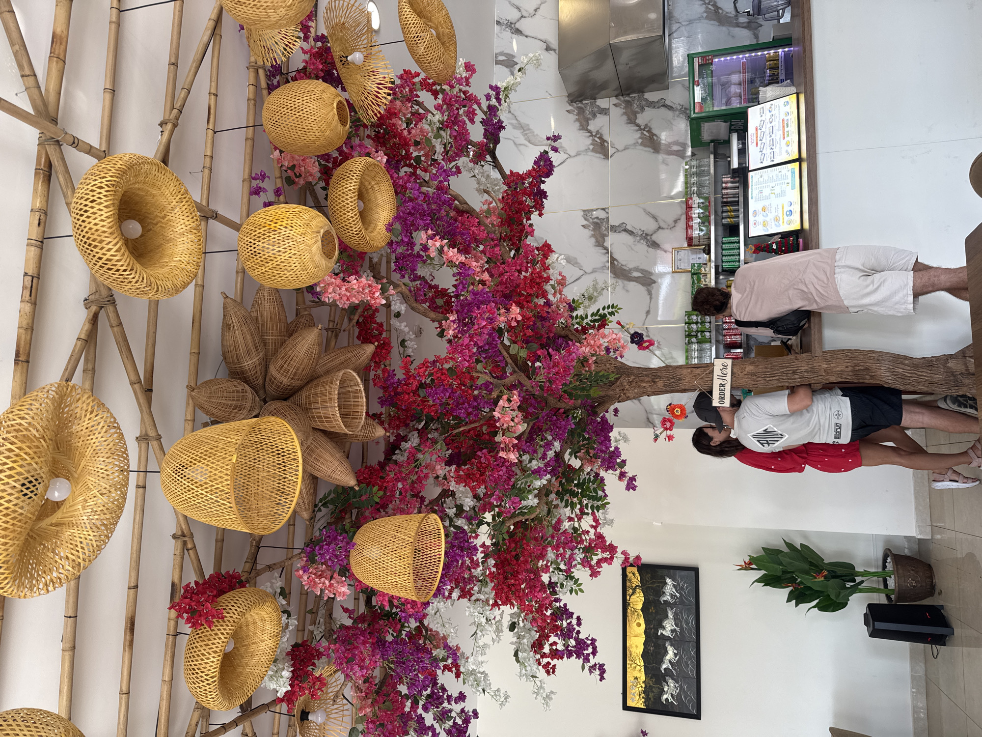 The image captures the interior of a food establishment, likely in Đà Nẵng, Vietnam, based on design elements. A decorative installation dominates the left side of the frame, featuring bamboo poles arranged vertically and horizontally, adorned with woven rattan or bamboo lampshades. Colorful artificial flowers in red, pink, purple, and white are interspersed with the bamboo structure.
To the right, people stand near a counter. One individual, wearing a pink shirt and white shorts, appears to be looking at a menu displayed above the counter. Another person, wearing a gray t-shirt, stands next to them. A signboard that says "Order Here" is stuck on the side of a tree.
Behind the counter, a beverage cooler is visible, stocked with cans and bottles. A framed picture hangs on the wall, depicting gold horses against a dark background. A potted plant stands near the picture.
The floor is light-colored tile. The walls have a white marble pattern and are a neutral white. The overall ambiance suggests a casual dining setting with a focus on decorative interior design.