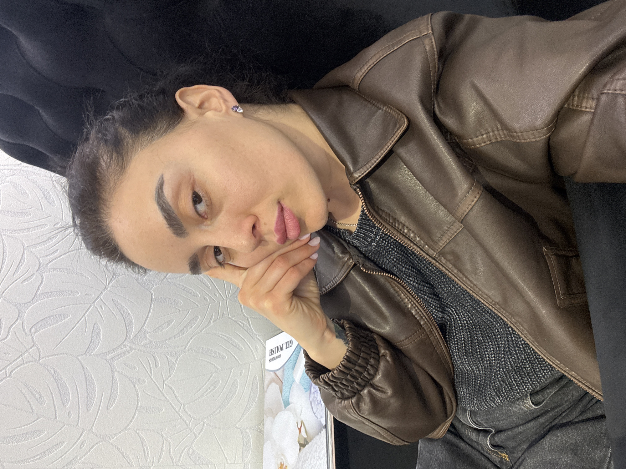A young woman is resting her head on her hand. She has dark hair pulled back, and wears a brown leather jacket over a gray sweater and dark jeans. She has earrings. The background includes a patterned wall, a dark sofa, and a poster advertising gel polish. The location is unknown and cannot be verified as Tbilisi, Georgia from the provided information.