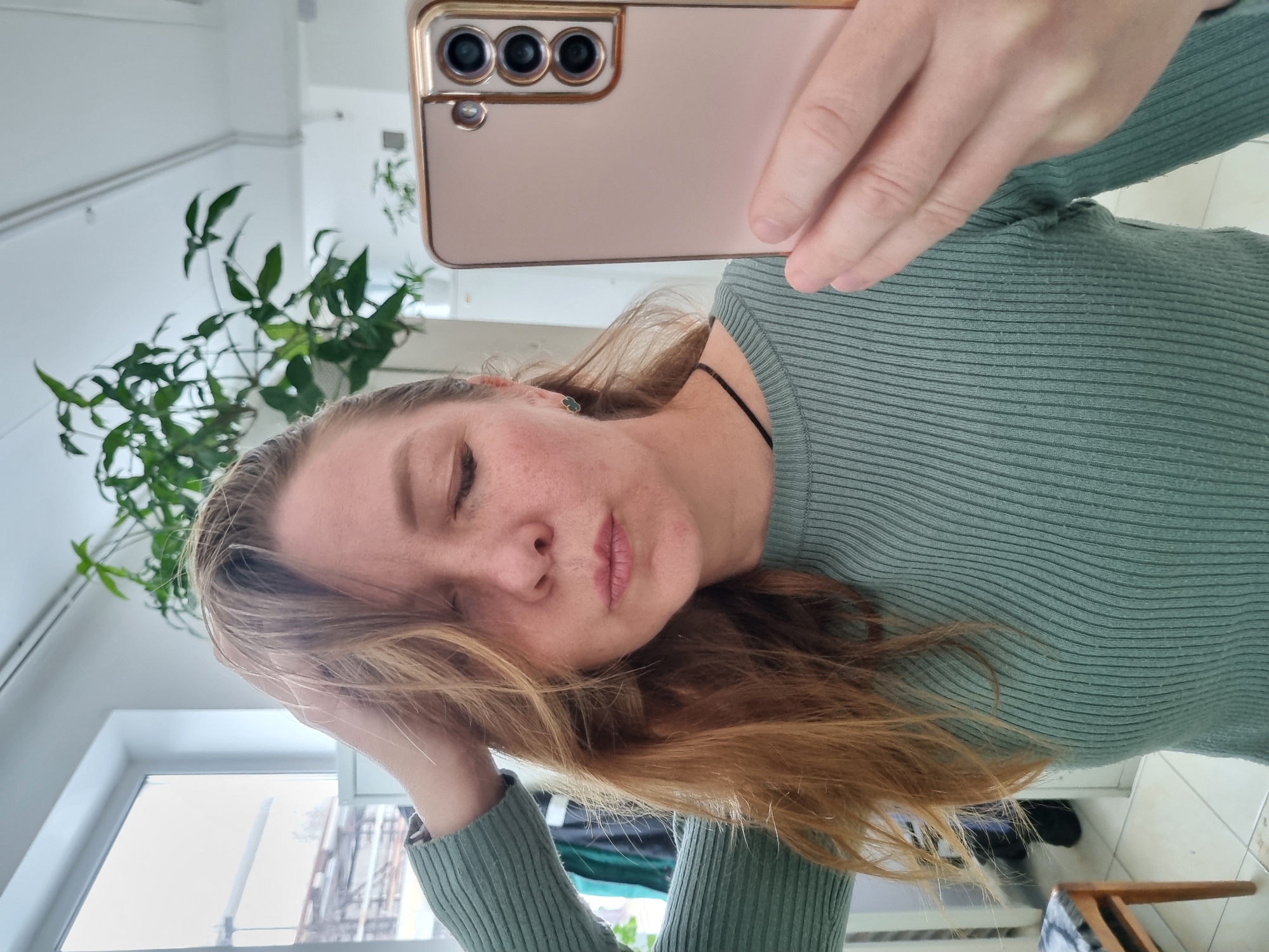 A woman with fair skin and freckles is taking a selfie indoors. She's wearing a ribbed, light-green long-sleeved shirt and a black choker necklace. Her long, light-brown hair is partially covering her face. She is holding a rose-gold smartphone with three rear cameras in her right hand, positioning it for the selfie. The background includes a window with an overcast sky and buildings visible outside, as well as a green potted plant. The location is not discernible as Kemul, Russia.