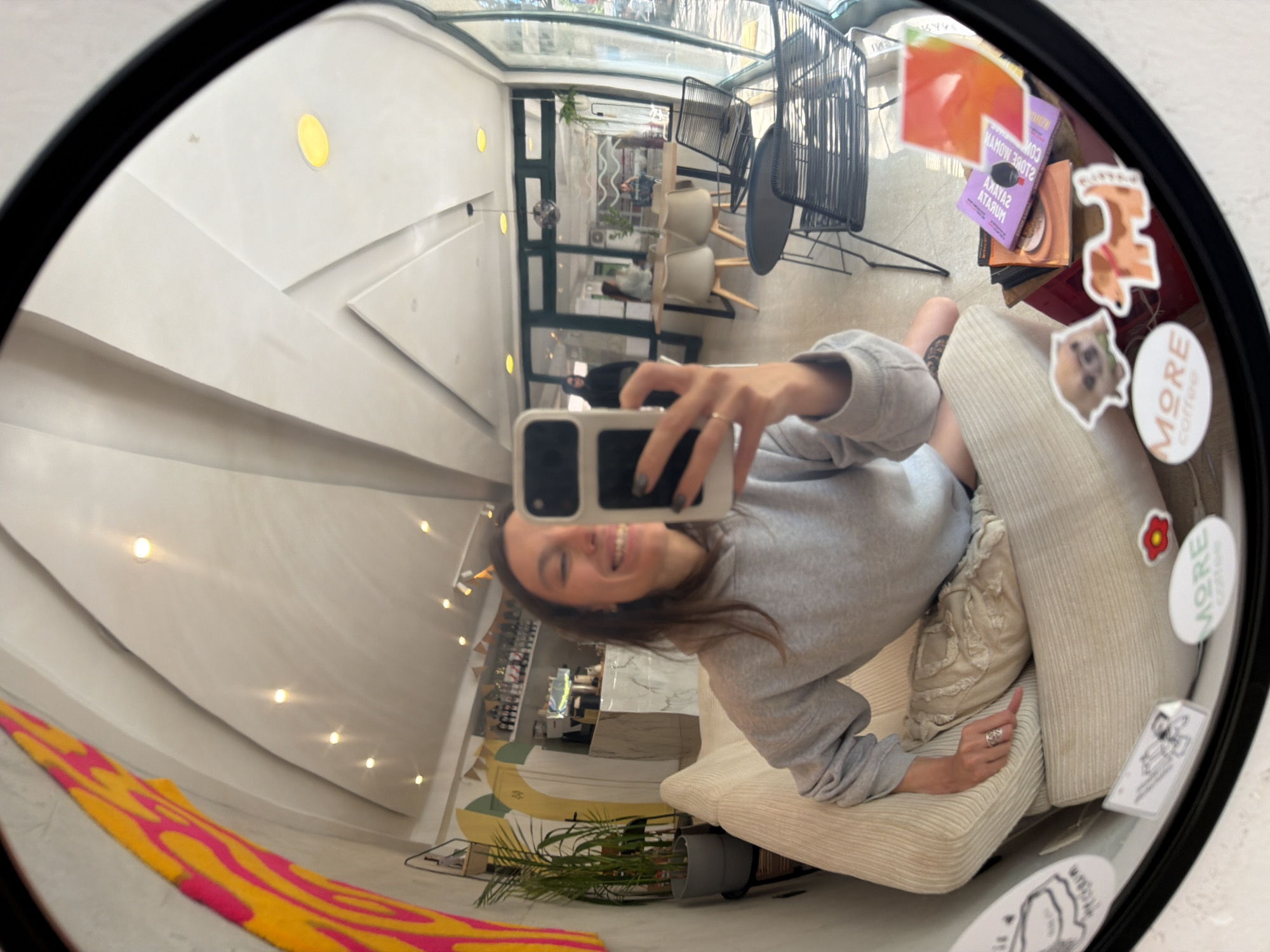 A mirror selfie shows a person with long brown hair smiling and holding up a mobile phone. She is wearing a grey sweatshirt and sitting on a light-colored couch. The mirror is round and has stickers on it. The reflection reveals part of a building interior, including chairs, tables, plants, and a yellow-and-pink design element. Based on the distinct interior design and a sticker that says "More Coffee," the location appears to be the "More Coffee" cafe in Budva, Montenegro.