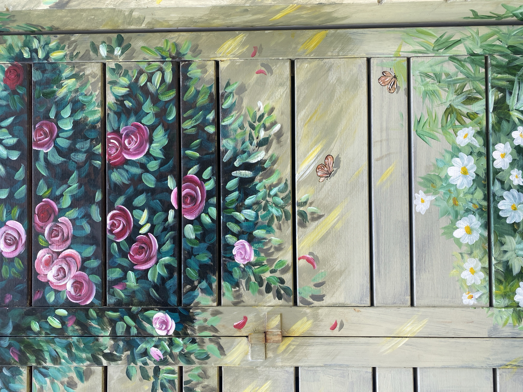 The image is a close-up, full-frame shot of a wooden surface, likely a shutter or a door, adorned with a vibrant, hand-painted mural. The artwork depicts a garden scene with a prominent display of pink roses and green foliage, interspersed with delicate white flowers and a few small butterflies. The style of painting is impressionistic, with visible brushstrokes and a rich texture.
The upper portion of the surface features dark wooden slats painted with lush green leaves and clusters of pink roses in various stages of bloom. The roses are depicted with detailed petals, ranging in hue from deep fuchsia to lighter pinks. The leaves are rendered in a variety of green tones, creating a sense of depth and volume.
As the mural progresses downwards, the background color transitions to lighter, more muted tones, suggestive of natural wood. Here, the flora becomes sparser, featuring more light pink roses and scattered red rose petals. Further down, small white flowers with yellow centers emerge from a bed of green leaves, accompanied by the depiction of slender bamboo-like stalks. Two small, brown butterflies with intricate wing patterns are also visible, one on a lighter-toned slat and another further down on the white flowers.
The wooden slats themselves are horizontally oriented, with visible grooves between them, indicating a louvered or paneled structure. A vertical seam runs down the left side of the frame, suggesting this is part of a larger object. There are subtle hints of yellow and white paint strokes, possibly representing sunlight or light reflections, especially on the left side of the mural. A metal latch or hinge is partially visible on the left side, connecting two sections of the wooden surface, adding a touch of realism to the painted scene.
While the image does not explicitly show any elements that directly place it in Da Nang, Vietnam, the style of folk art and the use of such decorative murals on outdoor surfaces are common in various urban and coastal areas of Vietnam, including Da Nang, often seen on shops, cafes, or residential properties to enhance their aesthetic appeal. The bright colors and floral motifs are in keeping with a tropical or sub-tropical setting.
