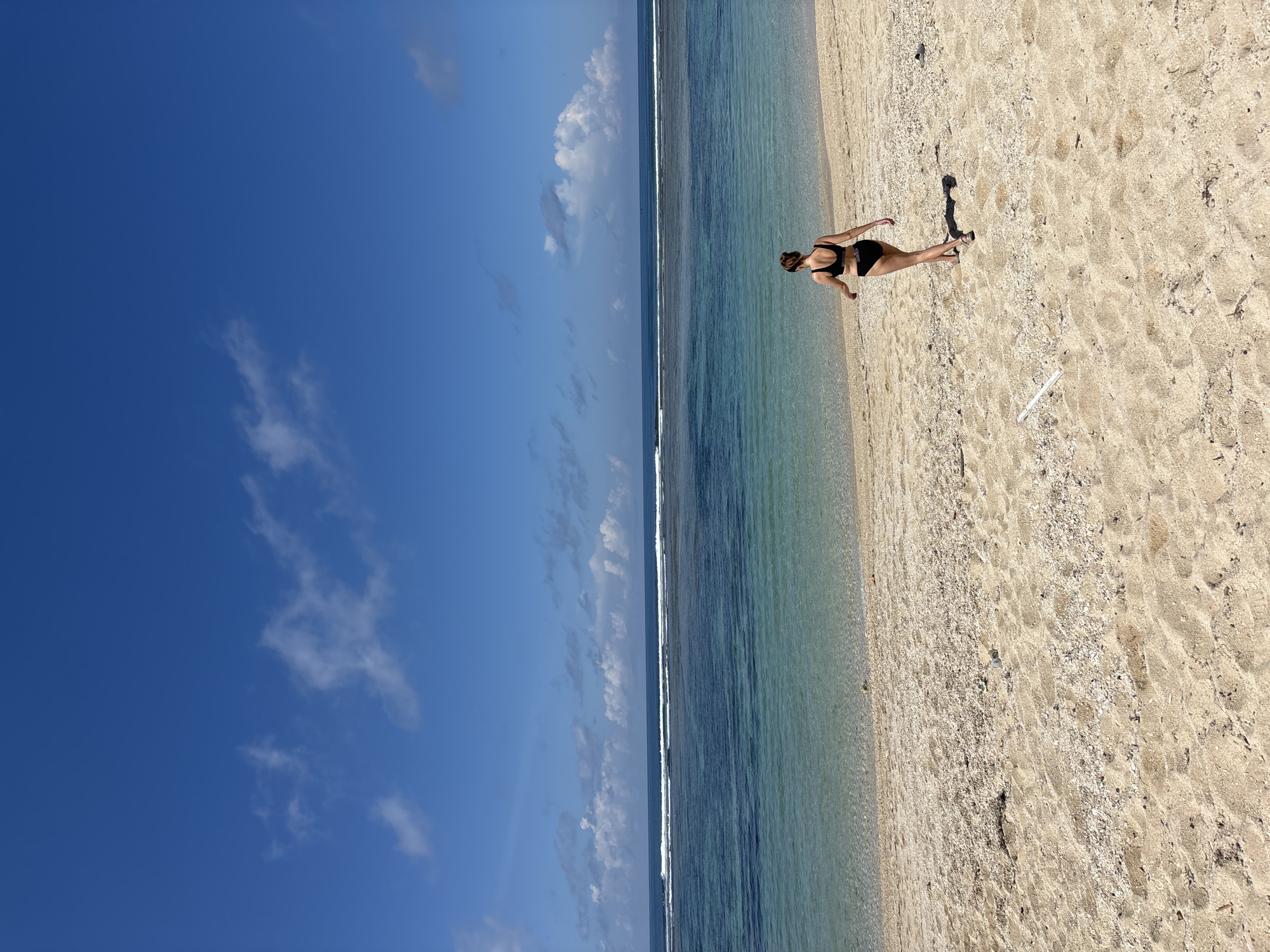 A photograph taken in Kutuh, Indonesia, shows a beach scene under a blue sky with scattered clouds. The ocean runs horizontally across the middle of the frame, transitioning from a light blue near the shore to a darker blue further out. On the right side of the image, a sandy beach covers the bottom half, appearing light tan and dotted with small shells. A person wearing a black sports bra and shorts walks on the sand, leaving footprints. Their shadow is visible on the beach next to them. The horizon line is mostly level, with the sky and ocean filling the left side of the image.