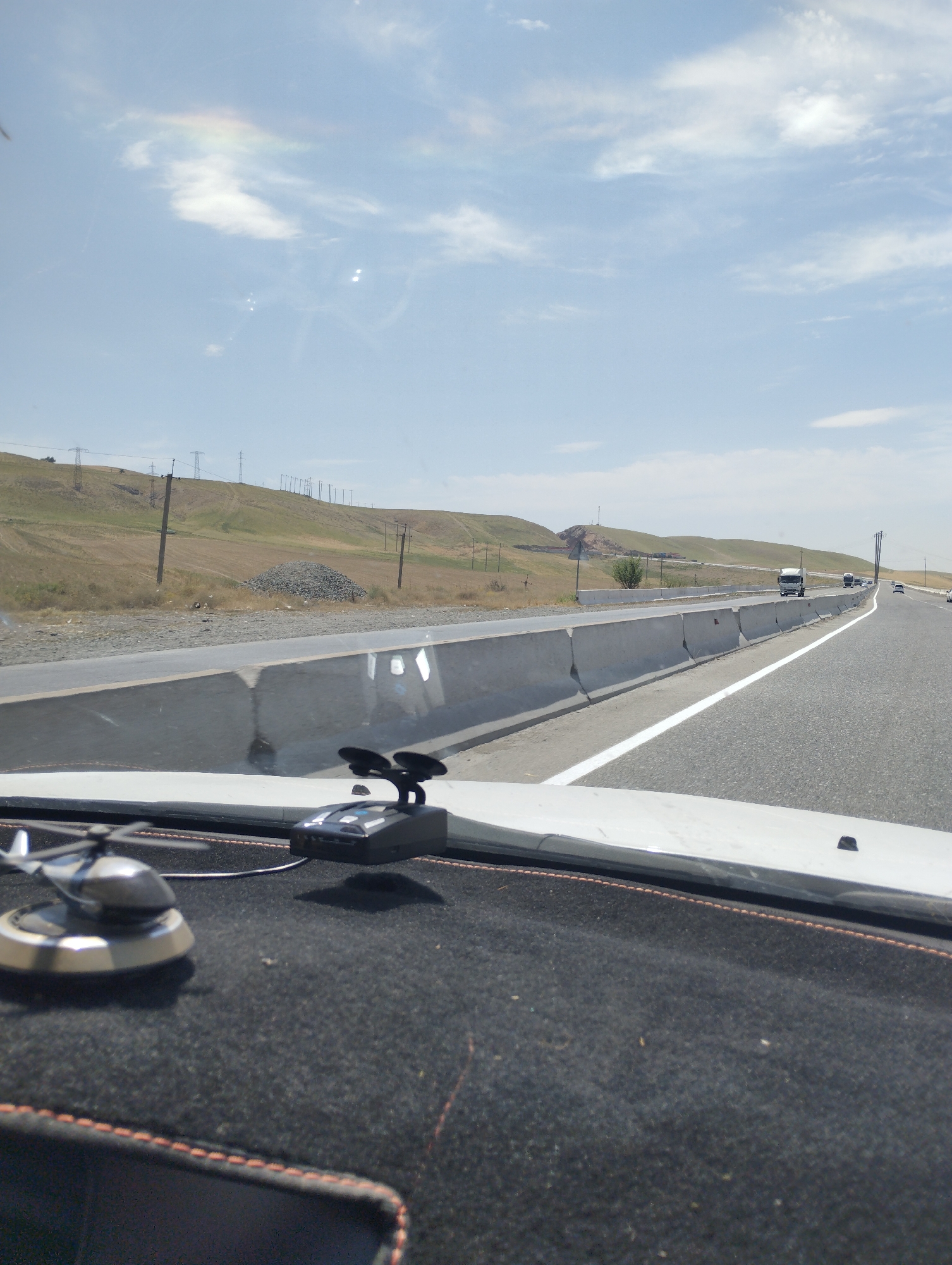 The image, taken from inside a vehicle, shows a view of a highway in Sangzar, Uzbekistan, on a clear, sunny day. The road ahead is paved and has a white lane marker. On the left side of the highway, there is a concrete barrier and beyond that, a dry, grassy hillside with power lines. The sky is blue with scattered white clouds, and a slight rainbow-like effect is visible in the upper left portion of the sky. In the foreground, on the dashboard of the vehicle, there is a small silver helicopter ornament and a black radar detector. Several vehicles, including a white truck, are visible on the highway in the distance. The overall scene depicts a typical highway landscape with elements of infrastructure and natural surroundings.