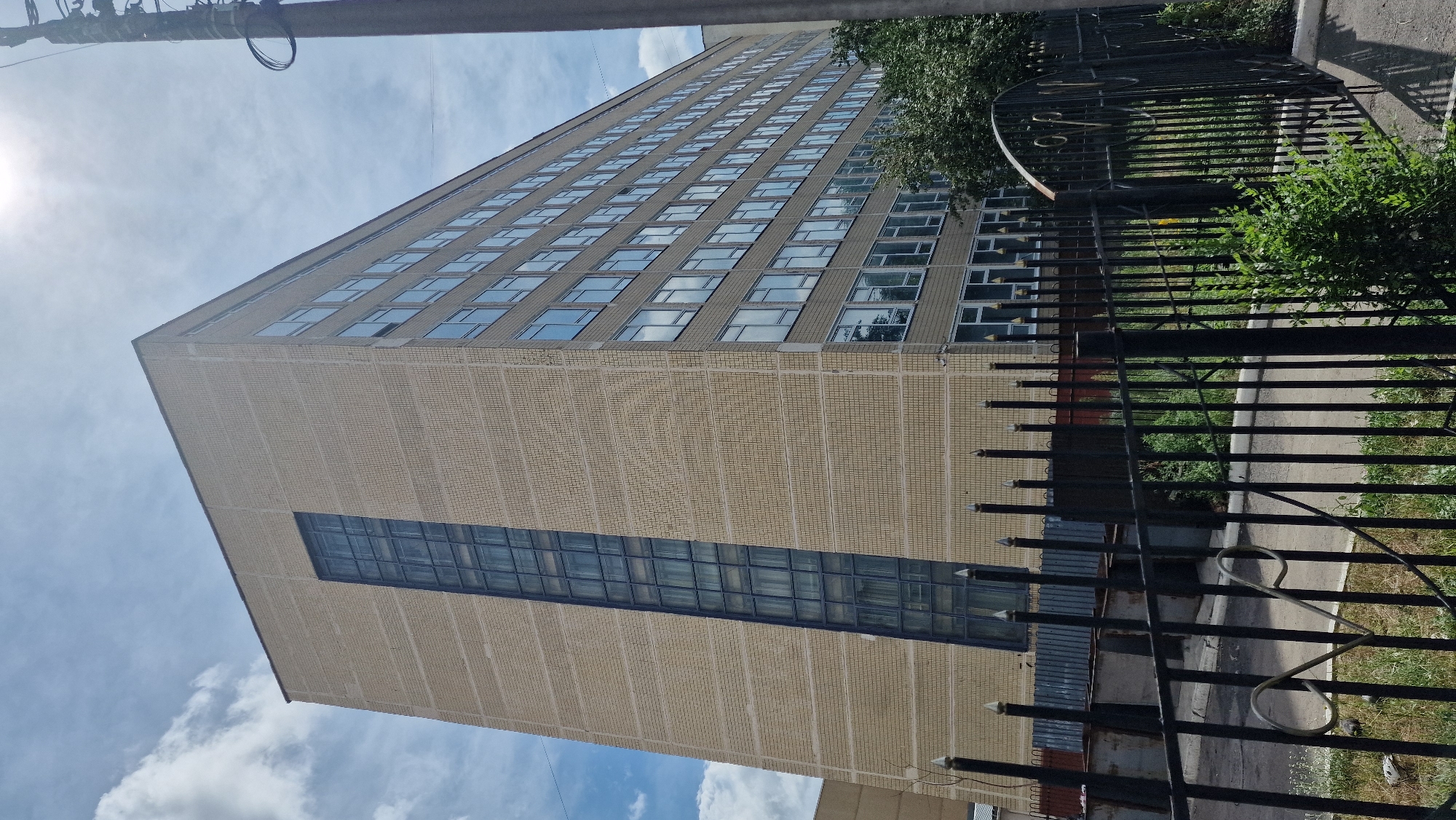 An exterior, eye-level, medium shot shows a light brown brick building with multiple floors and many windows. The building is partially obscured by a black metal fence in the foreground. The sky above is blue with scattered white clouds and a bright sun. The building has a large, vertical section of dark-tinted windows running up its side, and rows of smaller, rectangular windows along its facade. The fence is composed of vertical metal bars with decorative elements and pointed finials. Behind the fence, some dark-colored trash receptacles and green foliage are visible. A utility pole with wires is present on the right side of the image. The ground appears to be paved in some areas and grassy in others.