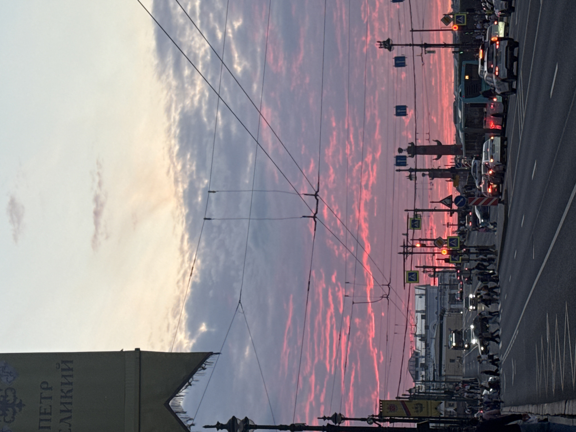 The image depicts a bustling city street scene under a dramatic sky, likely at sunset or sunrise. The sky is a prominent feature, showcasing a striking gradient from light blue-gray and white clouds on the left to an intense, fiery pink and red on the right, illuminated by the low-lying sun.
A wide asphalt road, marked with lane lines, stretches into the distance, filled with traffic. Numerous cars, including sedans and a bus, are present, with many displaying illuminated headlights or taillights. On the right side of the road, a group of pedestrians can be seen crossing at a designated crosswalk.
Overhead, a complex web of electrical wires, likely for trolleybuses or trams, crisscrosses the sky, supported by utility poles that line the street. Various road signs are visible, including blue pedestrian crossing signs, directional arrows, and red traffic lights. Buildings with European architectural styles are discernible in the background.
On the far left, a banner or flag is partially visible, bearing Cyrillic text. The legible portion reads "ПЕТР ВЕЛИКИЙ" (PETER THE GREAT), a strong indicator that the location is Saint Petersburg, Russia. The combined effect of the vivid sky, busy traffic, and illuminated street elements creates a dynamic and captivating urban landscape.