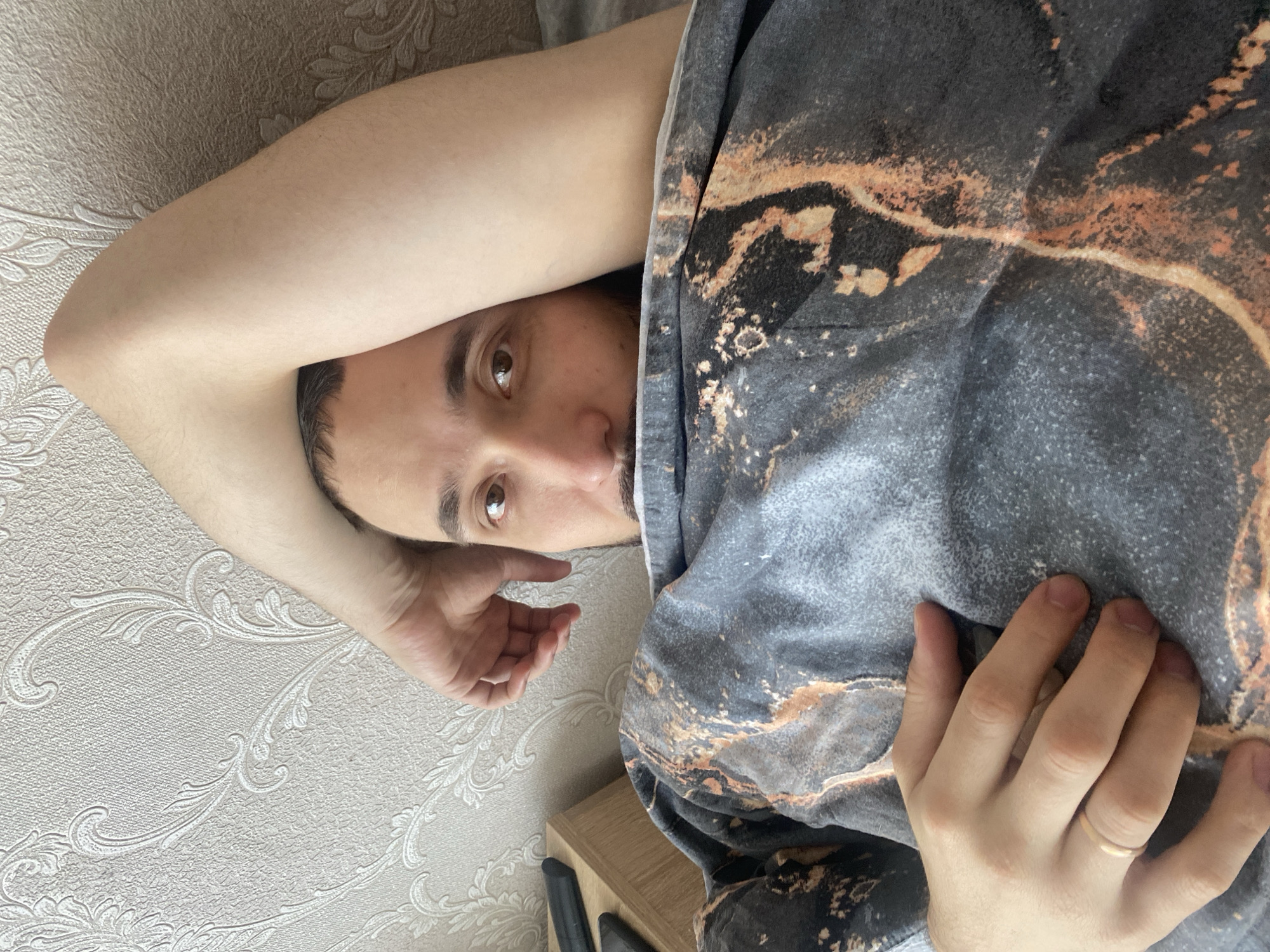 A man is lying down, face turned toward the camera. His left arm is bent and positioned above his head, resting on a textured, light-colored wall. His right arm is extended and slightly bent, with his hand resting on top of a patterned fabric. The fabric is a mix of grey, black, and brown colors with a marble-like pattern. A small portion of a light wood surface and two black objects are visible beneath the fabric. The man has short dark hair, a mustache, and light brown eyes. The image was taken indoors. There is no information to indicate that this photo was taken in the city of Chaikovsky, Russia.