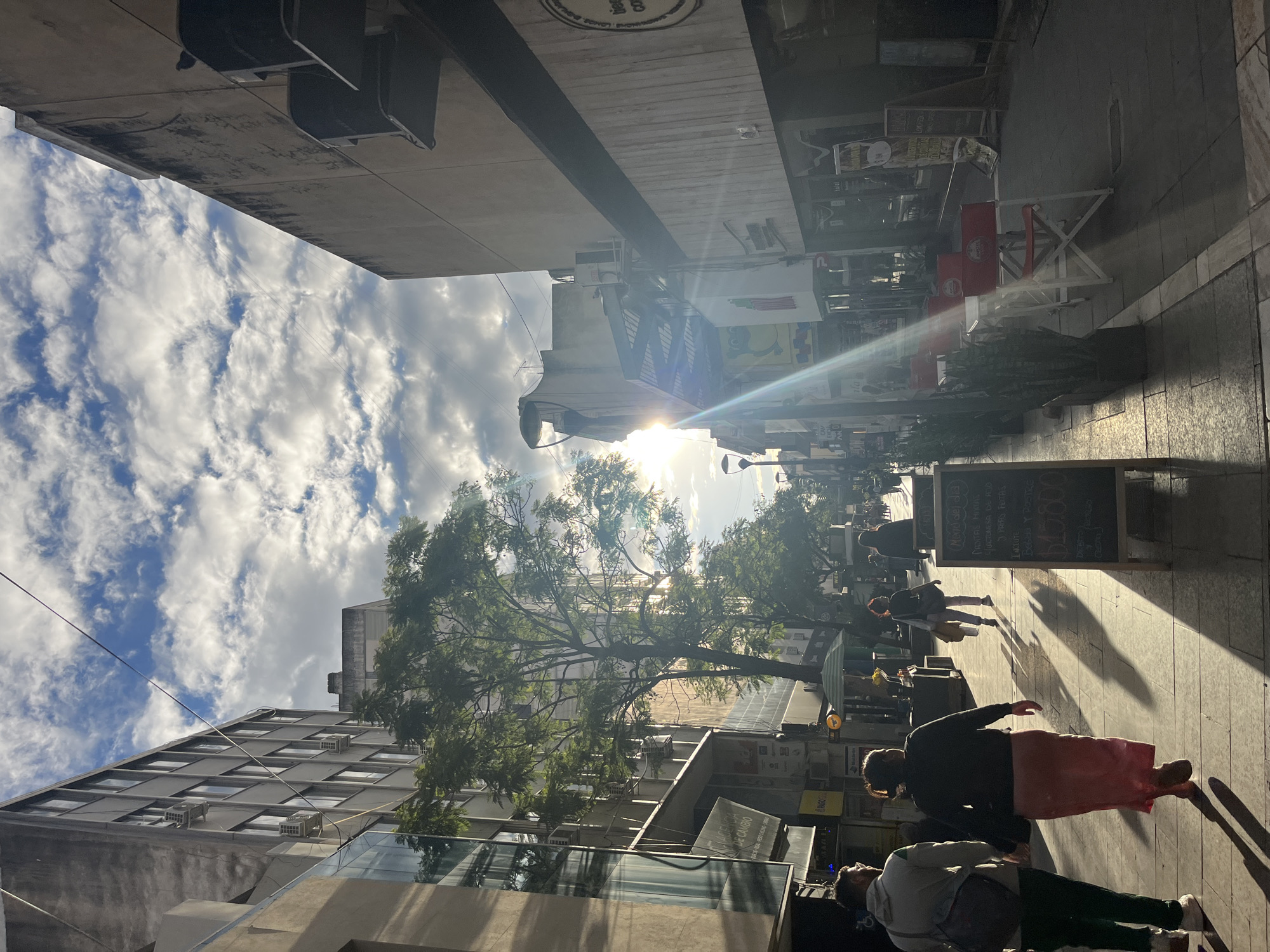 This image captures a vibrant, sunlit city street, likely in Cordoba, Argentina, given the provided context and visible Spanish text. The scene is viewed from a slightly elevated perspective, looking down a street or pedestrian area flanked by buildings.
The time of day appears to be either early morning or late afternoon, as the sun is low in the sky, creating a strong lens flare and casting long, distinct shadows across the paved sidewalk. The sky above is partially cloudy, with scattered white clouds against a blue background.
Numerous pedestrians are seen walking and standing on the sidewalk, engaging in typical urban activities. In the immediate foreground, a person in a pink skirt and another in a grey hoodie and green pants are visible. Further along, more individuals can be seen near commercial establishments.
The street is lined with multi-story buildings housing various businesses. On the right side of the image, an outdoor cafe setup features red and white tables and chairs, some of which bear the "Amstel" beer logo. Two large chalkboards stand on the sidewalk, displaying menu information in Spanish. One sign advertises a "Menú del día" (Menu of the day) that "INCLUYE Bebida Y POSTRE" (Includes Drink and Dessert) with options such as "PAPAS FRITAS" (French Fries) and a price of "$1.500". Another prominent sign indicates an "AGENCIA DE CAMBIO" (Money Exchange Agency).
A large, verdant tree with extensive foliage stands in the middle of the street, its branches extending upwards, providing a touch of nature amidst the urban setting. Overhead, power lines crisscross the frame. The overall atmosphere is one of a lively, everyday city scene.